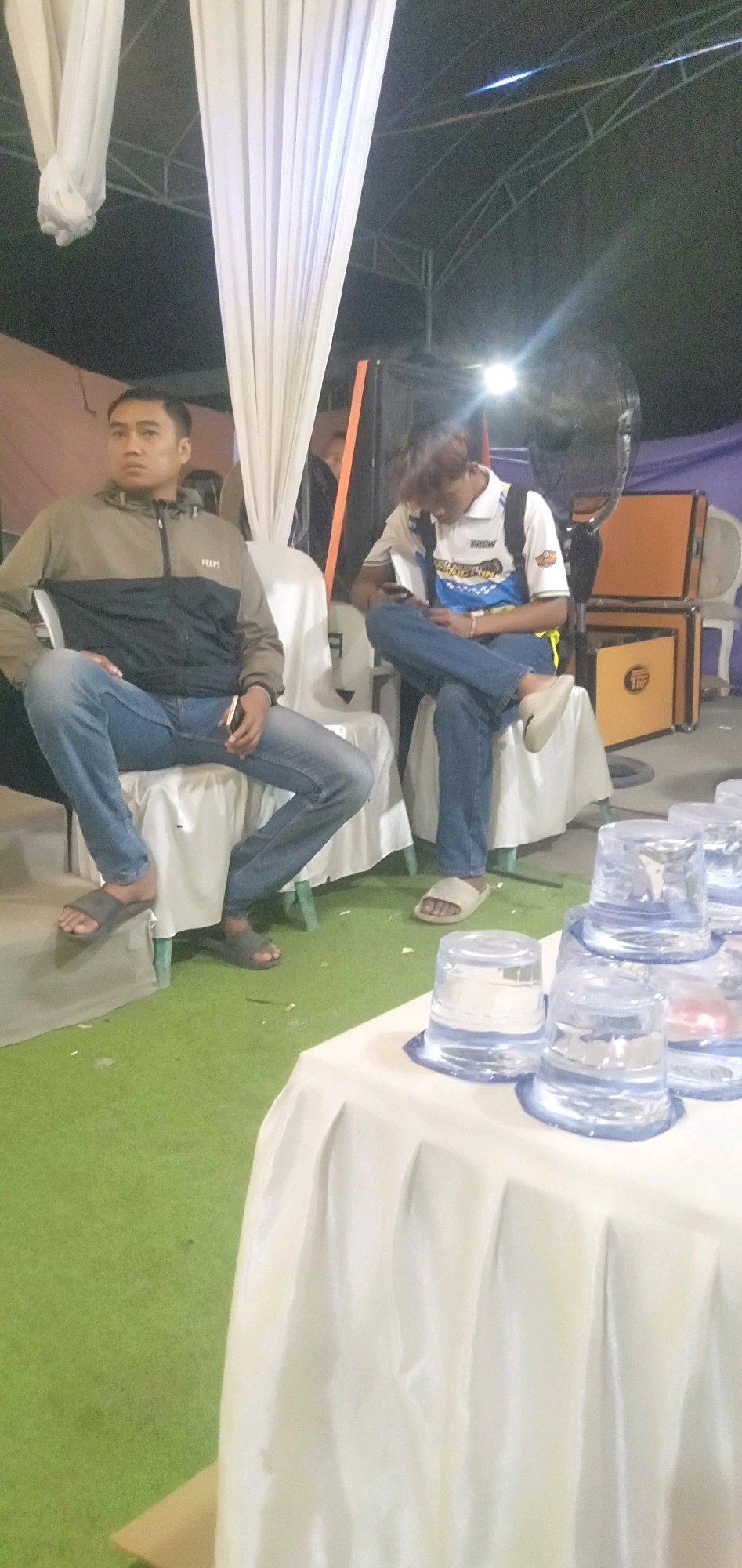 Two young men are seated on chairs covered with white fabric. The man on the left, wearing a brown and black jacket and blue jeans, is looking directly at the camera. The man on the right, in a white and blue shirt and blue jeans, is looking down at his phone. They are positioned on a green carpeted area, possibly an outdoor event space, with white drapery hanging in the background. To the right, there are stacks of clear plastic cups filled with water on a table covered with a white tablecloth. The environment suggests a casual gathering or event. The scene appears to be captured at night, as indicated by the artificial lighting and dark sky. The location is identified as Plemahan, Indonesia.