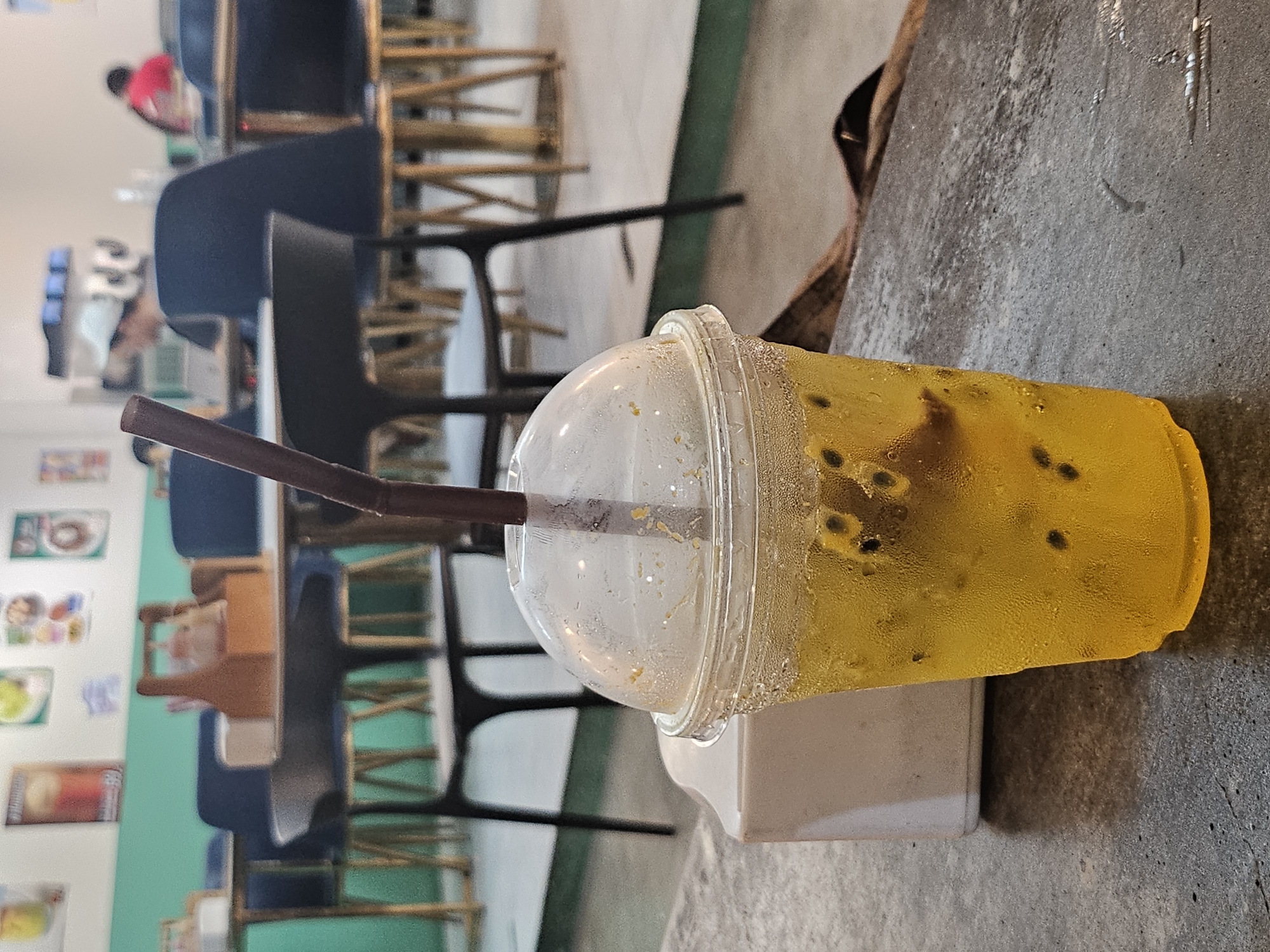 The image shows a close-up of a plastic cup of passion fruit juice with pulp and seeds. The cup is sweating and sits on a textured gray surface, possibly a table. In the background, out of focus, is the interior of a restaurant or cafe in Ratsada, Thailand. There are several tables and chairs, with a mint green wall visible behind them. A wooden condiment caddy is on one table. A few patrons are present but are blurred, including a person wearing a white cap and another in a red shirt. The overall atmosphere suggests a casual dining environment.
