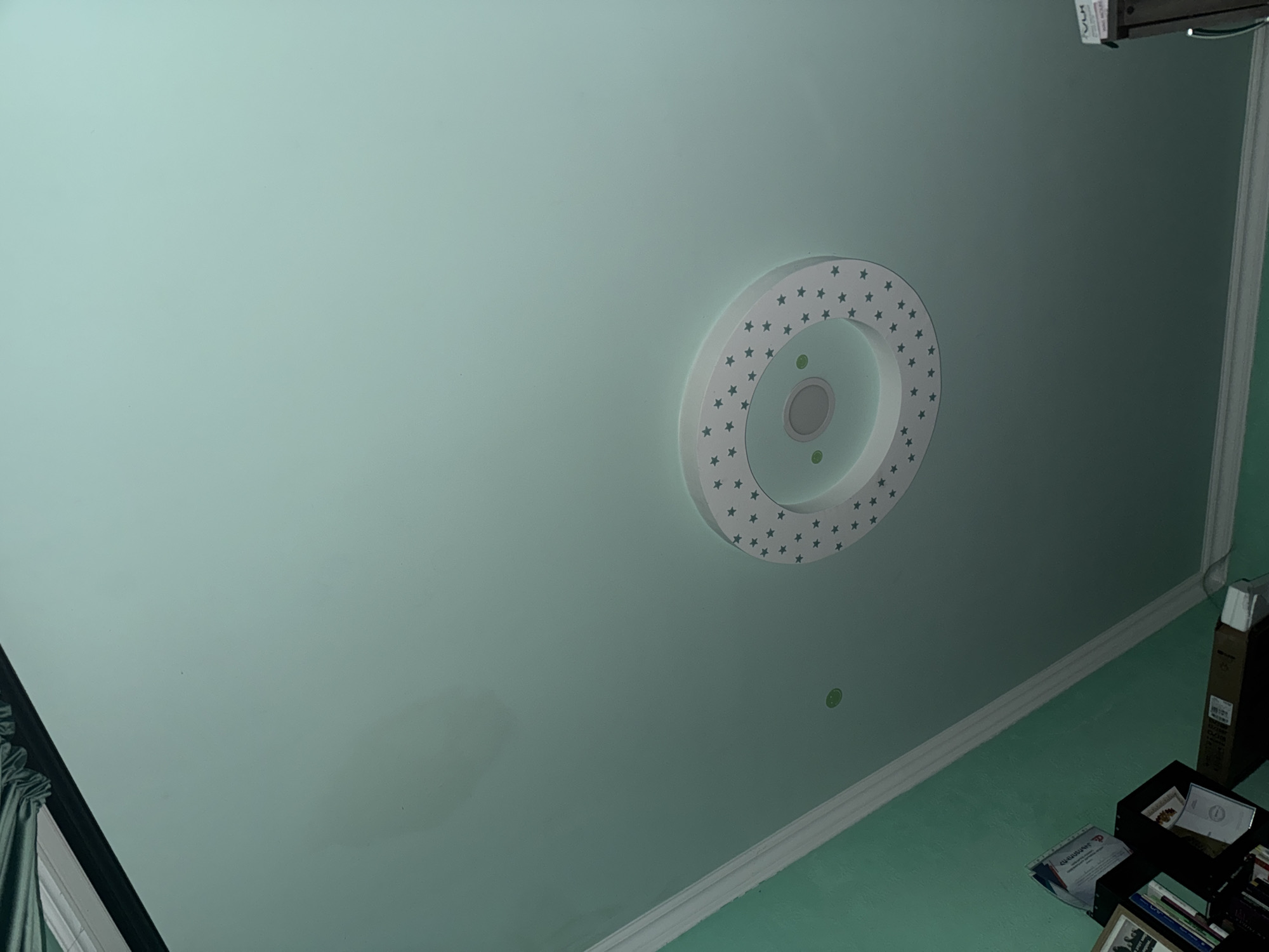 The image shows a view of a pale green ceiling. At the center of the frame is a circular light fixture with a ring of star-shaped cutouts. The lighting is soft and diffused. In the lower-right corner, there's a glimpse of a shelf with various objects, including boxes, books, and papers. Parts of the wall and a curtain are visible along the edges of the image. The overall setting appears to be indoors, likely in a residential building. The location in the city of Yerevan, Armenia is not possible to determine from the content of this image.
