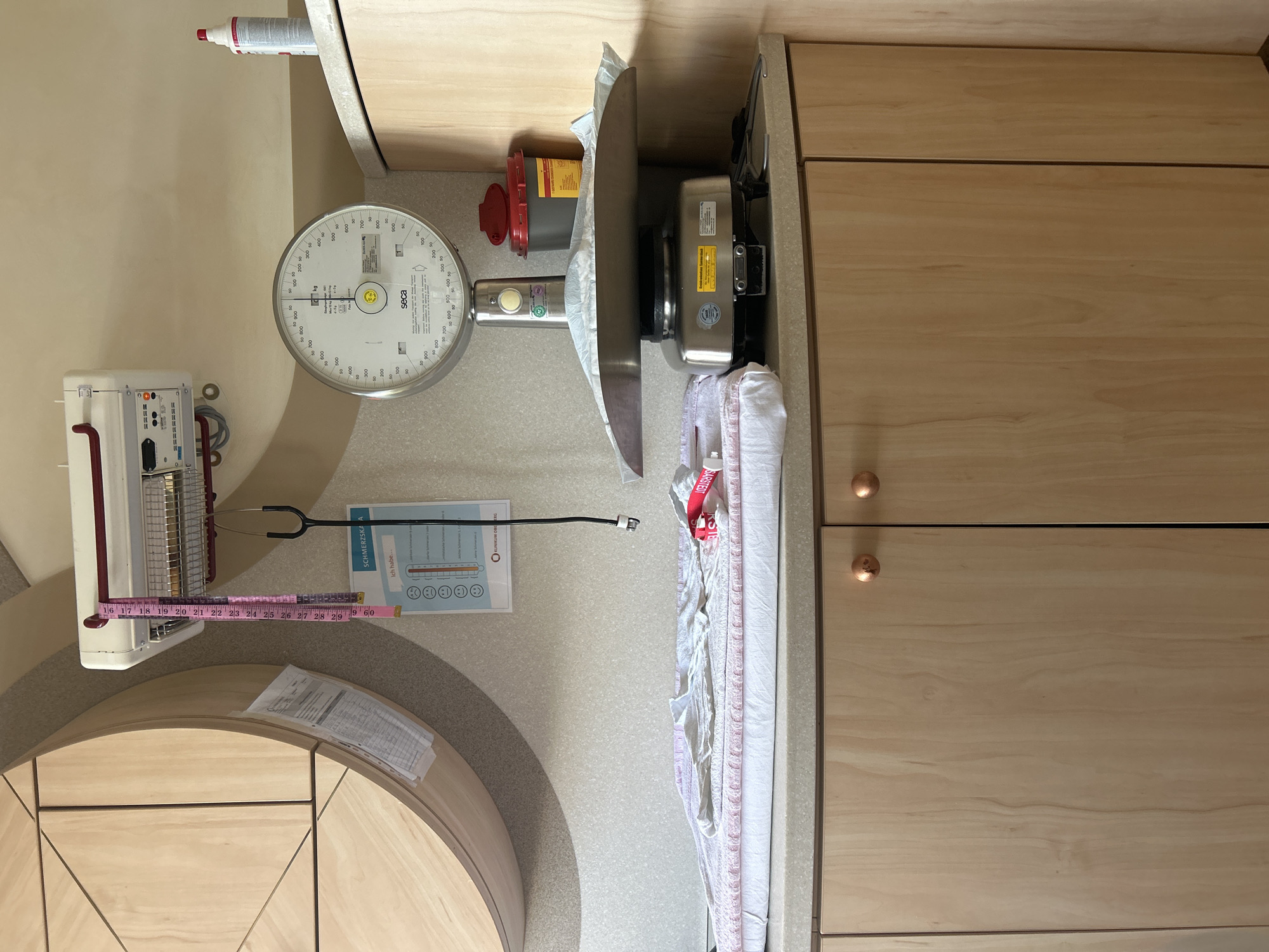 The image depicts a medical examination room, likely in a hospital or clinic, featuring a baby scale, a changing table with linens, and various medical instruments and supplies. The scale, a SECA model, is positioned on a countertop above wooden cabinetry. A pink measuring tape hangs from a wall-mounted unit. A stethoscope is also visible, along with a hazardous waste container and a bottle of disinfectant. A chart displaying pain scales is affixed to the wall. The room's lighting suggests it is an interior space. The scene is tidy and organized, consistent with a healthcare setting. Based on the presence of the "KLINIKUM OBERBERG" logo on the pain chart, the location is the Oberberg Klinikum in Gummersbach, Germany.