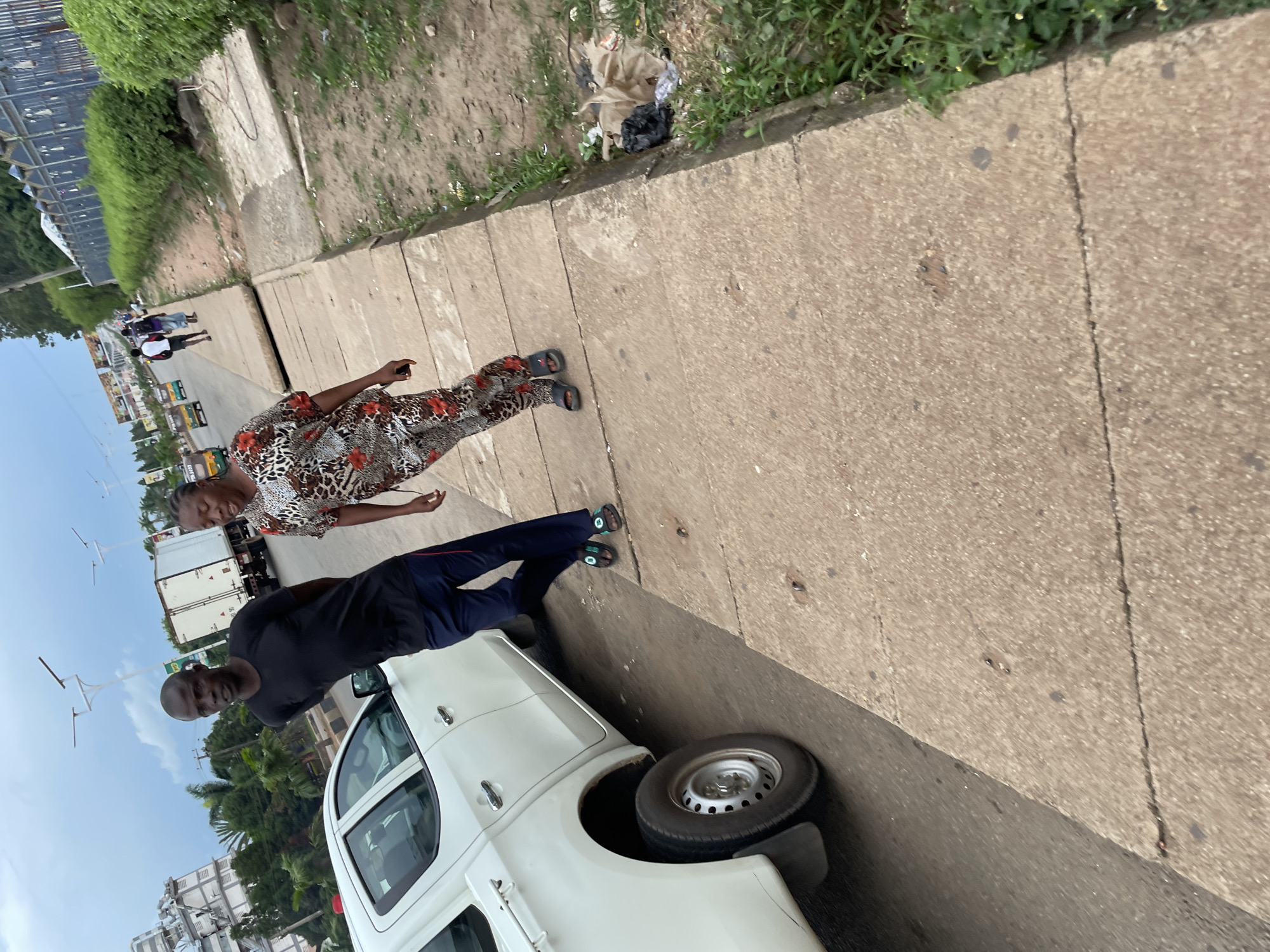 A man and a woman walk on a concrete sidewalk next to a white pickup truck on a cloudy day in Jos, Nigeria. The man is wearing a black t-shirt and blue pants. The woman is wearing a patterned shirt and pants. In the background, there are trucks, cars, and buildings. There is trash on the side of the road.