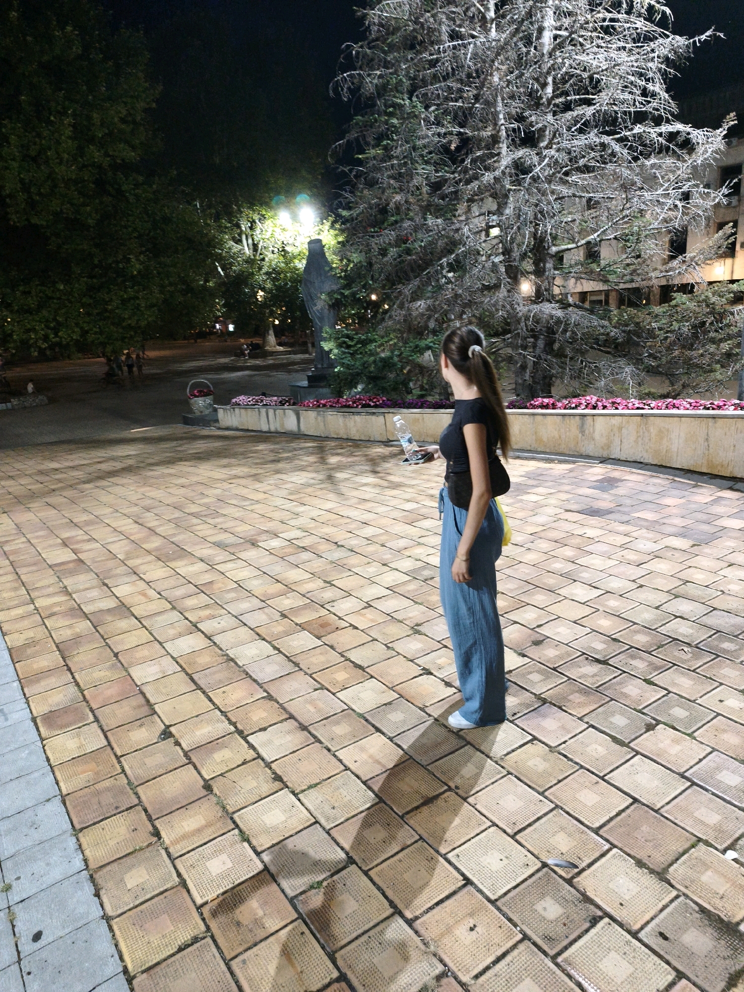 A young woman with a ponytail, wearing a black top and blue pants, stands on a paved plaza at night. She is holding a water bottle and looking towards a statue in the background. The plaza is lit by streetlights, illuminating the tiled ground and surrounding trees. In the background, there are buildings, a statue, and more trees. The scene appears to be a public space in the city of Lovech, Bulgaria.