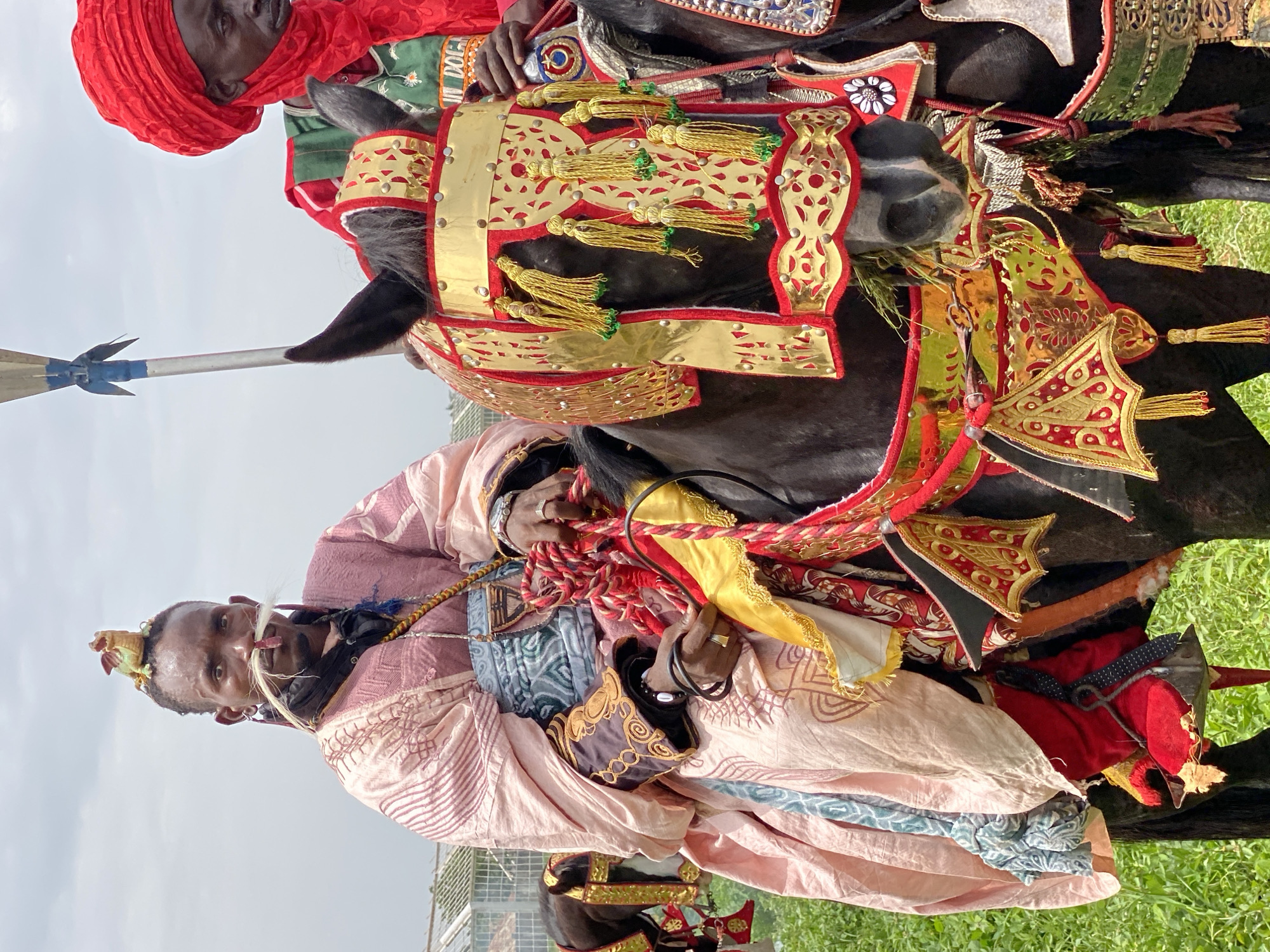 The image depicts two men on horseback, likely captured in Kafanchan A, Nigeria, given the prompt. Both horses are adorned with elaborate, traditional ornamentation, including gilded and colorful fabrics. The rider closest to the viewer is wearing a light pink and patterned robe and possesses distinctive facial decorations, including white elements near the nose and ornate headwear. The second rider, behind and slightly above, is wearing a red turban. One rider holds the reins of the horse and displays jewelry on his fingers. The background shows green vegetation and a cloudy sky.