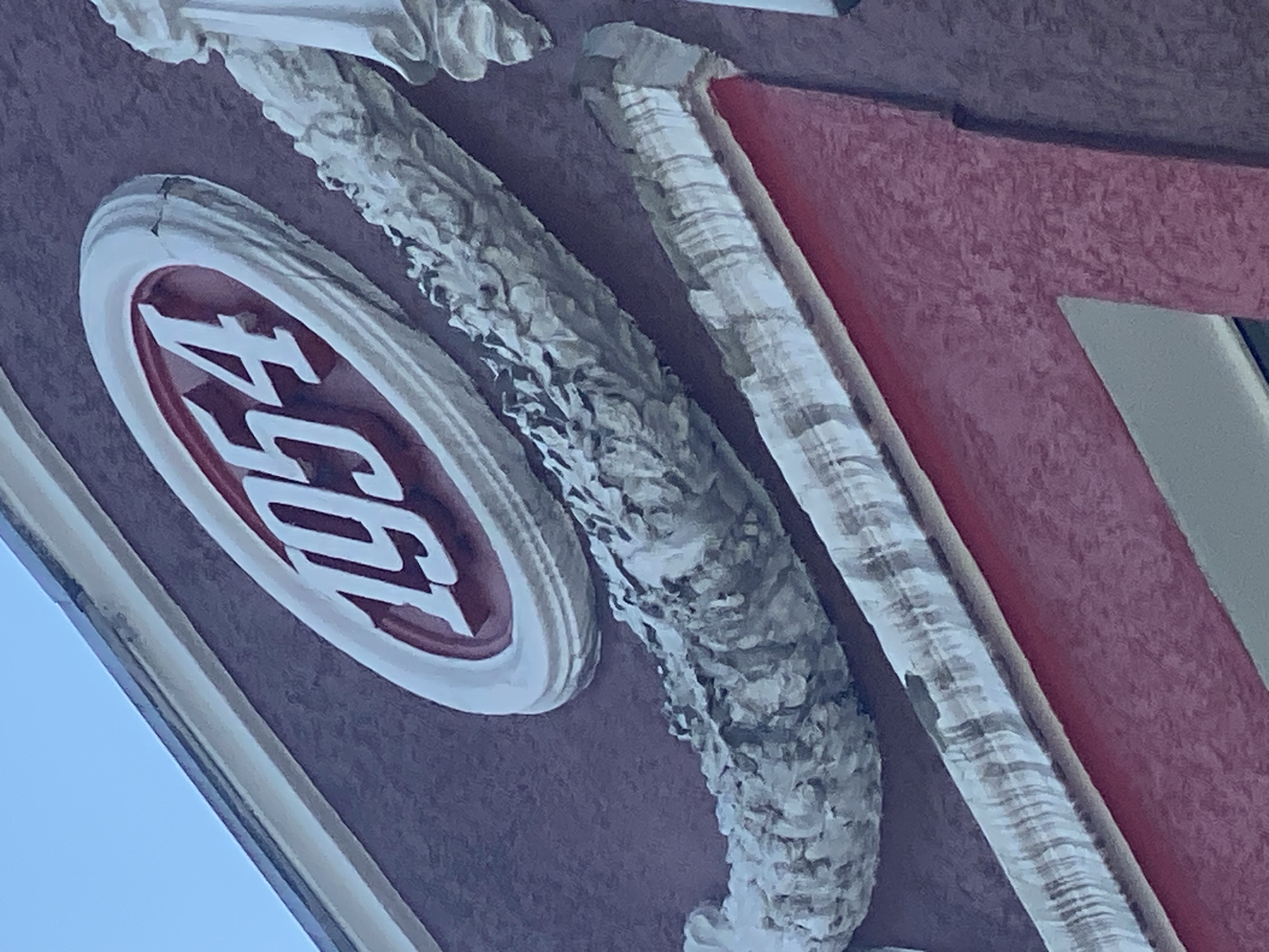 The image shows a close-up view of an oval-shaped emblem on a dark red building facade. The emblem contains the white numbers "1954" in a bold, serif font, with a red background within the oval. The emblem is bordered by a white, textured ring and appears to be made of plaster or a similar material. Below and to the right of the emblem, there is ornate decorative molding, also appearing to be made of plaster, in a pale, off-white color. This molding has a textured, branch-like or floral pattern and is layered. The building facade itself has a slightly textured finish. At the bottom of the image, a bright red architectural element is visible, possibly part of a window or doorway. The sky, a light blue, is visible in the upper left corner. The image is taken from a low angle, looking up at the building. This is a detailed architectural photograph focused on decorative elements and the building's date marker. There are no people or actions depicted in the image. The location is identified as Novokuznetsk, Russia.