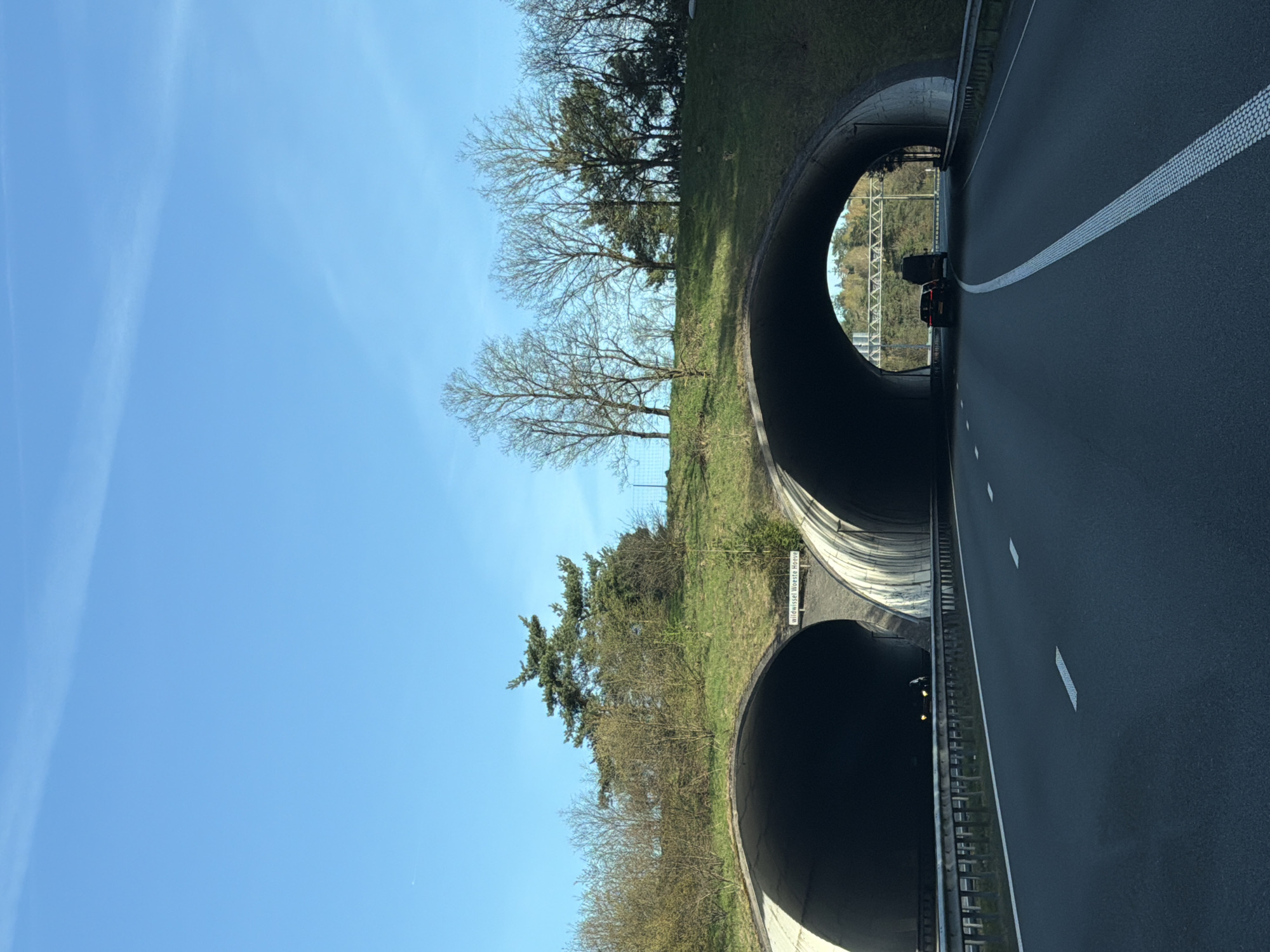 The image depicts a section of highway passing through a "wildwissel Woeste Hoeve" wildlife crossing tunnel in Beekbergen, Netherlands. Two tunnels are visible side-by-side, constructed through a grassy hill. Trees line the top of the hill and surround the tunnel entrances. A clear blue sky is above. The highway is asphalt, marked with solid white lines. There is a car visible within the upper tunnel.