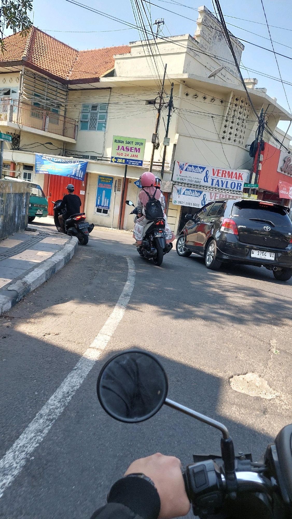 The image shows a street scene in Sidoarjo, Indonesia, with several motorcycles and a car. A person is riding a motorcycle in the foreground, with their hand on the handlebar and a side mirror visible. In the middle ground, two other motorcycles are visible, one carrying a passenger wearing a pink helmet. A black car is also present on the right side of the road. In the background, a building with a sign that reads "RUMAH SAKIT UMUM JASEM IGD 24 JAM" is visible, indicating a general hospital with 24-hour emergency services. The environment appears to be a typical urban street with buildings, power lines, and a paved road.