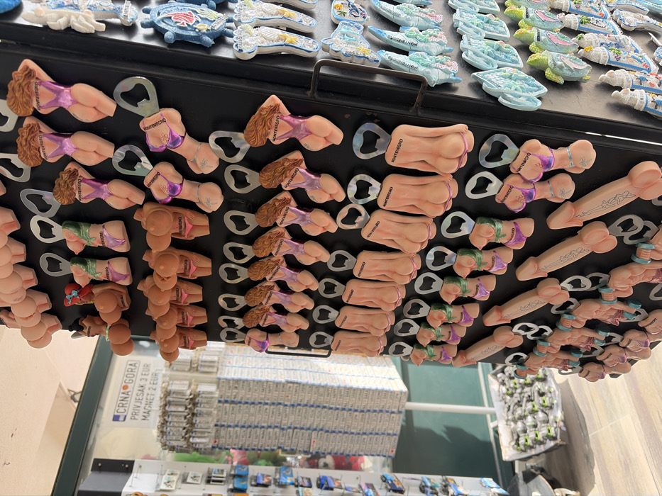 This image captures a close-up view of a souvenir display within an open-fronted shop or stall, located in Budva, Montenegro. The primary focus is a black display board vertically aligned on the right, showcasing numerous novelty bottle openers. These bottle openers predominantly feature anatomically suggestive designs, including rows of light-pink human buttocks, female torsos in various-colored bikinis (purple, green, orange), and phallic shapes, many inscribed with "MONTENEGRO."
To the right of this board, another section of the display holds more conventional souvenir magnets, depicting sea turtles, fish, and miniature architectural forms, primarily in shades of blue, white, and light green.
In the background on the left, additional souvenir merchandise is visible. A white sign with blue text reads "CRNA GORA" (Montenegro) and advertises "PRIVJESAK 3 EUR / MAGNET 2 EUR" (Keychain 3 EUR / Magnet 2 EUR). Below this sign, numerous small, rectangular magnets or keychains are arranged on a shelf. At the bottom left, a bin or cart contains a large quantity of small, circular metallic objects, likely more keychains or magnets. The floor appears to be light-colored tile or pavement, and the scene is brightly lit, suggesting an outdoor or semi-outdoor commercial setting. No individuals are visible in the image.