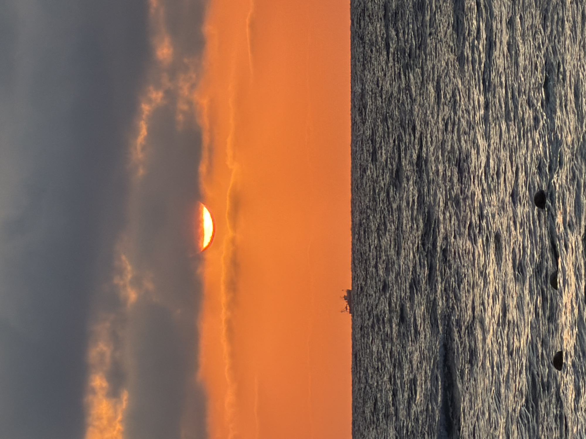 The image depicts a sunset over the Mediterranean Sea, likely taken from the coast of Tel-Aviv, Israel. The sky is a vibrant gradient of orange and yellow, with dark gray clouds forming a band across the upper portion. A portion of the sun is visible peeking through the clouds, casting a warm glow. In the middle ground, a small fishing boat is visible on the horizon. The foreground shows the dark, choppy waters of the sea, with two dark buoys floating on the surface. The overall scene is serene and picturesque, capturing the beauty of a coastal sunset.