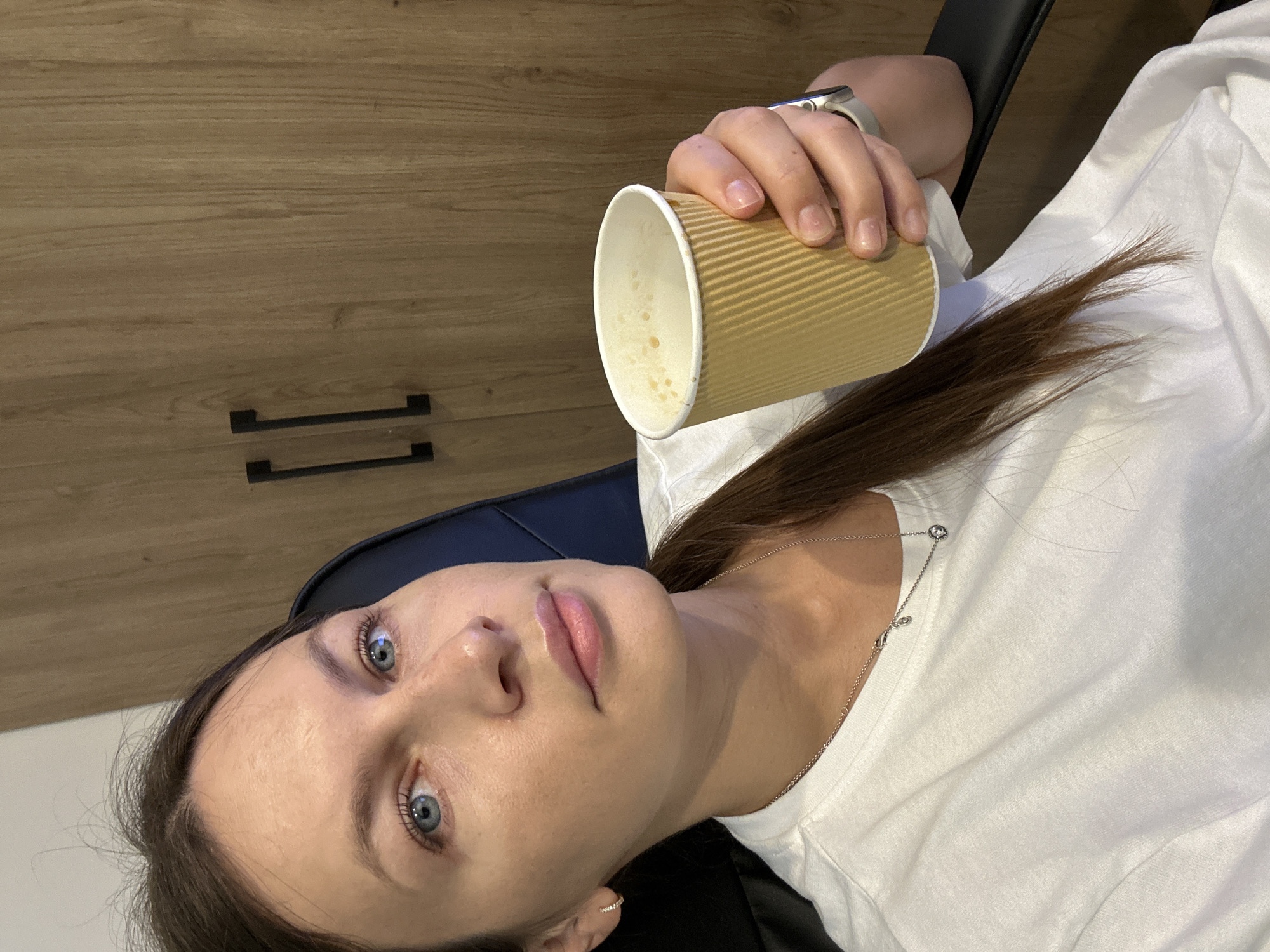 A young woman with blue eyes and brown hair tied in a ponytail sits in a black office chair, holding a disposable coffee cup in her right hand. She is wearing a white t-shirt and a silver necklace with a small pendant. The background features a light brown wooden cabinet with two black handles. The woman appears to be in an indoor environment, possibly an office or a home. The scene is well-lit, with the focus on the woman and her coffee cup. There is no visible indication of the city of Boreti, Montenegro in the image.