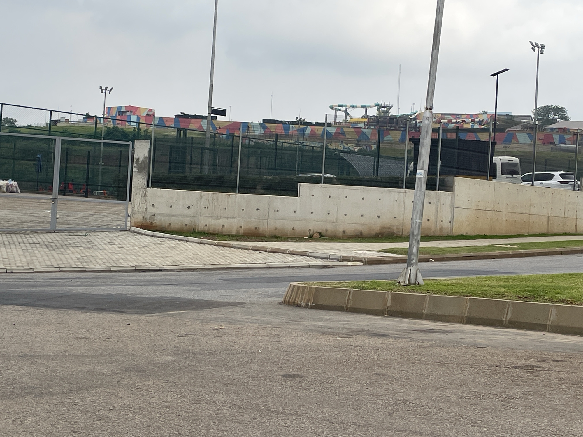 The image shows a view of the "Splash" water park in Abuja, Nigeria. In the foreground, there is an asphalt road and a paved walkway leading to a metal fence. Behind the fence, a concrete wall runs horizontally, separating the lower area from the upper part of the park. The upper area features colorful structures of the water park, including slides and tiered seating areas decorated with geometric patterns in red, blue, yellow, and green. Two white vehicles, a van and a car, are parked near the water park entrance. The sky is overcast.
