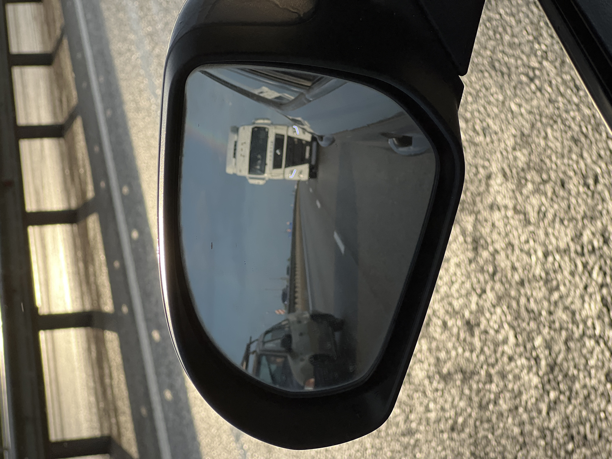 In the reflection of a car's side mirror, a white semi-trailer truck is visible driving on a highway. The truck is approaching a rainbow visible in the sky. In the left side of the mirror, a darker colored hatchback car with a roof rack is visible from the rear. The road surface appears wet and reflective, suggesting recent rain. The scene takes place during daylight, with the sun casting long shadows from a guardrail in the foreground. The location is Psekups, Russia.