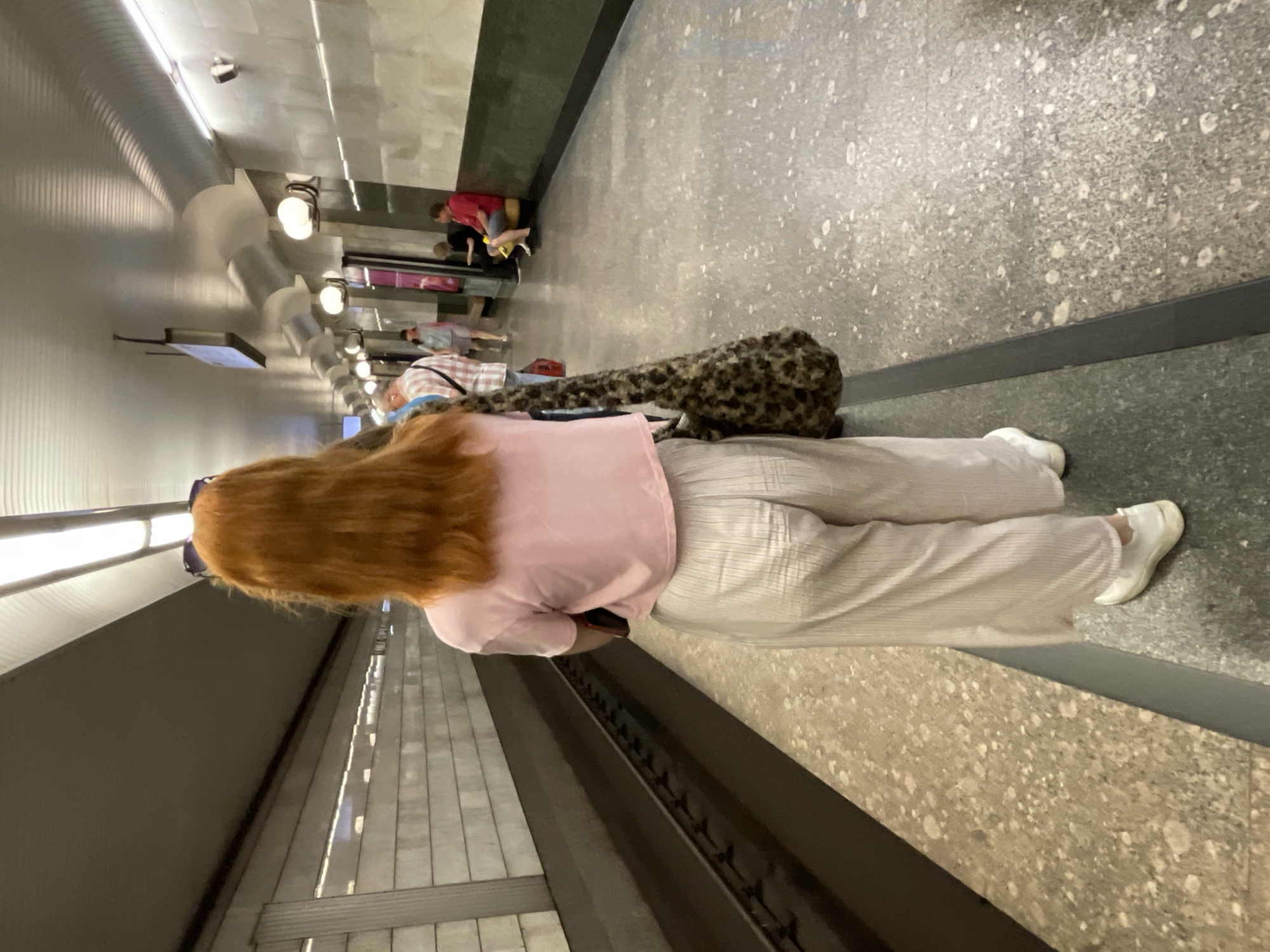 A woman with long, red hair and sunglasses on her head stands with her back to the camera, facing the subway tracks in a Moscow metro station. She is wearing a pale pink cropped t-shirt and loose-fitting, cream-colored striped pants. She carries a large, fuzzy, leopard-print bag over her right shoulder. The station platform is made of speckled grey and beige tiles, with a dark grey border. The walls of the station are clad in textured grey panels and lit by fluorescent lights and globe-shaped lamps. Several other people are visible further down the platform, some sitting on benches and others standing.