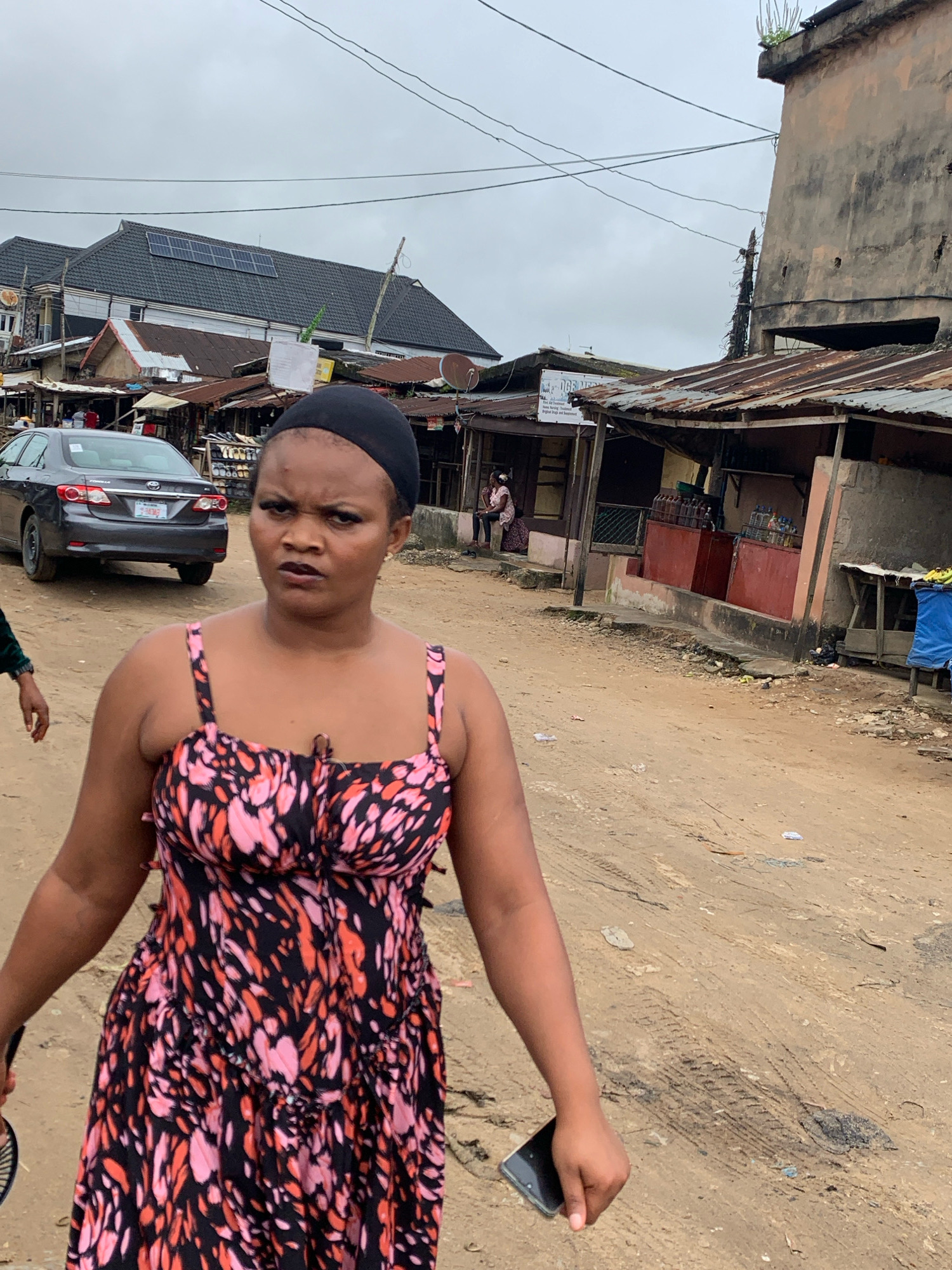 A woman wearing a black head wrap and a black, pink, and orange patterned dress is walking towards the camera on a dirt road in Umuode, Nigeria. She is holding a smartphone in her right hand. Behind her, a grey sedan is parked on the left side of the road. The street is lined with various buildings, some with corrugated metal roofs. To the right, there are stalls or shops with goods displayed. The sky is overcast.