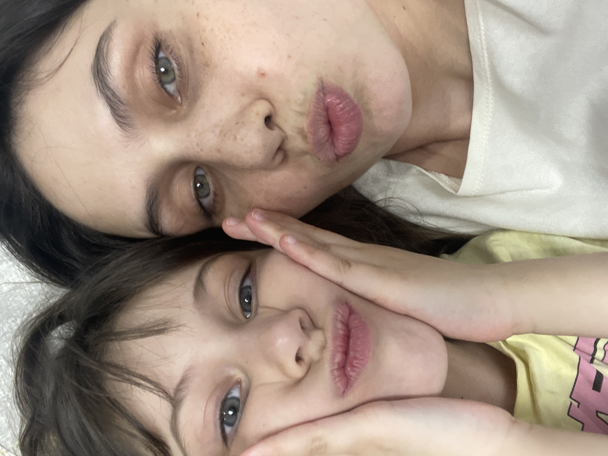 A close-up, intimate portrait, captured indoors in Izhevsk, Russia, features two females, likely a mother and child, posed affectionately. The image is tilted, showcasing their faces side-by-side, both looking towards the camera with playfully pursed lips in a "kiss" expression.
The adult, positioned above, has dark hair, light skin with visible freckles, prominent eyebrows, and striking green eyes. She wears a light-colored or white top. Her head gently rests upon the child's, conveying a sense of warmth and connection.
The child, in the lower part of the frame, also has dark hair and green eyes. She is wearing a light yellow top, which partially reveals a graphic with pink lettering. Her hands are playfully placed around her face, emphasizing her puckered lips.
The overall mood is tender and playful, capturing a moment of bonding. Both subjects share similar facial features, including eye color. The lighting is soft and even, highlighting their expressions. There is no visible text within the image.