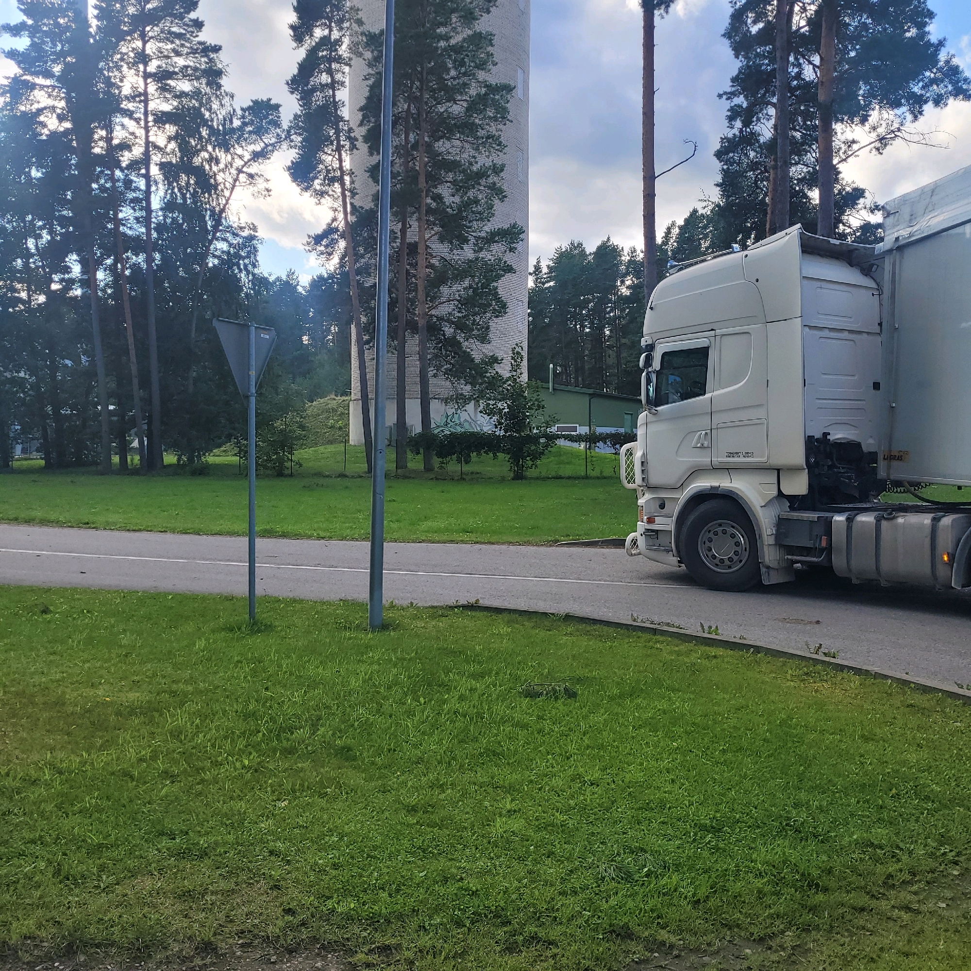 The image captures a white semi-trailer truck driving on a road in Jūrmala, Latvia. The truck is positioned on the right side of the frame, with its front cab visible, including headlights, grille, and wheels. A portion of the trailer is also visible, attached to the truck. The truck is on an asphalt road with a white line marking.
To the left of the road, there is a well-maintained grassy area. Further back, a tall, cylindrical white structure, possibly a water tower, stands amidst a backdrop of tall pine trees and other greenery. A grey triangular traffic sign on a pole is also visible in the grassy area, close to the road. The sky above is partly cloudy with some blue sky peeking through. The overall environment suggests an outdoor setting, likely in an industrial or semi-industrial area given the presence of the large truck and the water tower.