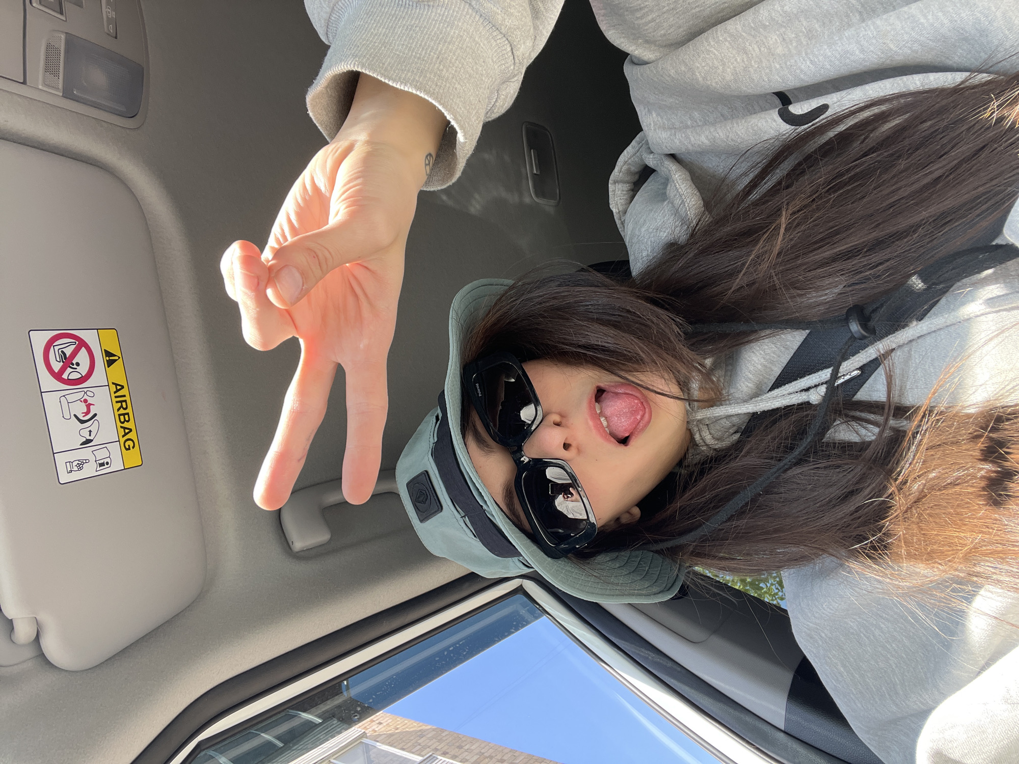 A playful, upside-down selfie is captured inside a car on a bright, sunny day. The primary subject is a person with long, dark brown hair, seen from a low angle, looking upwards towards the vehicle's ceiling. They are wearing a grey hooded sweatshirt, a light green or grey bucket hat, and black sunglasses, with the brand "Polaroid" faintly visible on the frame. The person is making a peace sign with their left hand, holding it close to the camera, and has their mouth open with their tongue sticking out, conveying a humorous and cheeky expression.
The car's interior features a light-colored headliner and a sun visor. On the sun visor, a yellow warning label prominently displays "AIRBAG" in bold text, accompanied by pictograms. These symbols include warnings against placing a rear-facing child seat in the front seat when an airbag is present, and a reminder to fasten seatbelts. A small, stylized tattoo, possibly a peace symbol, is visible on the wrist of the person's right arm, partially covered by the hoodie sleeve.
Bright sunlight streams into the car, illuminating the scene and casting shadows. Through the car window at the bottom of the frame, a clear blue sky is visible, along with a glimpse of a building or structure. The image conveys a casual, lighthearted mood captured with an unconventional, inverted perspective.