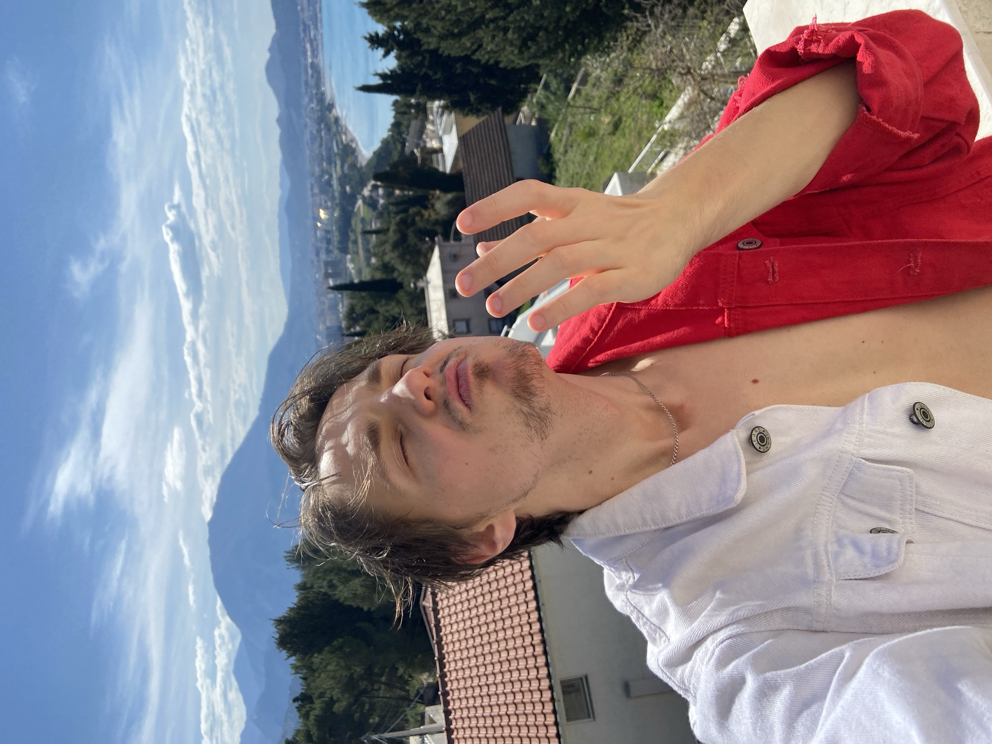 A man is taking a selfie outdoors. He has brown hair and stubble, and he is wearing a white denim jacket and a red shirt. He is facing upwards with his eyes closed and his lips pursed. His right hand is raised in front of his face. In the background is a landscape with mountains, trees, and buildings. The location appears to be Šušanj, Montenegro, based on the coastal view and architecture. The weather is sunny.