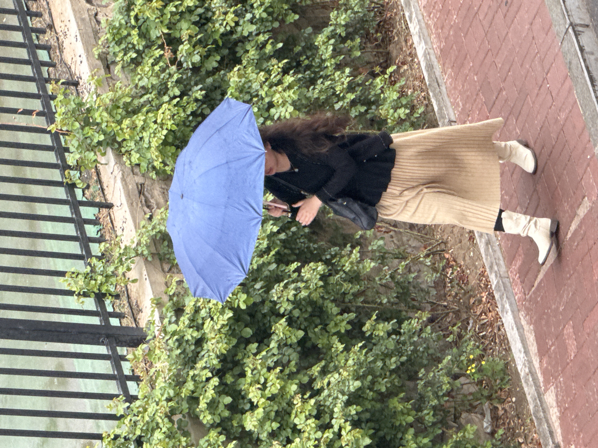 A woman with long, dark hair walks on a brick sidewalk in Zhongtun, China. She is holding a blue umbrella to shield herself from the rain. She is wearing a black jacket, a tan skirt, white boots, and black socks. The sidewalk is adjacent to a green shrubbery and a black metal fence.