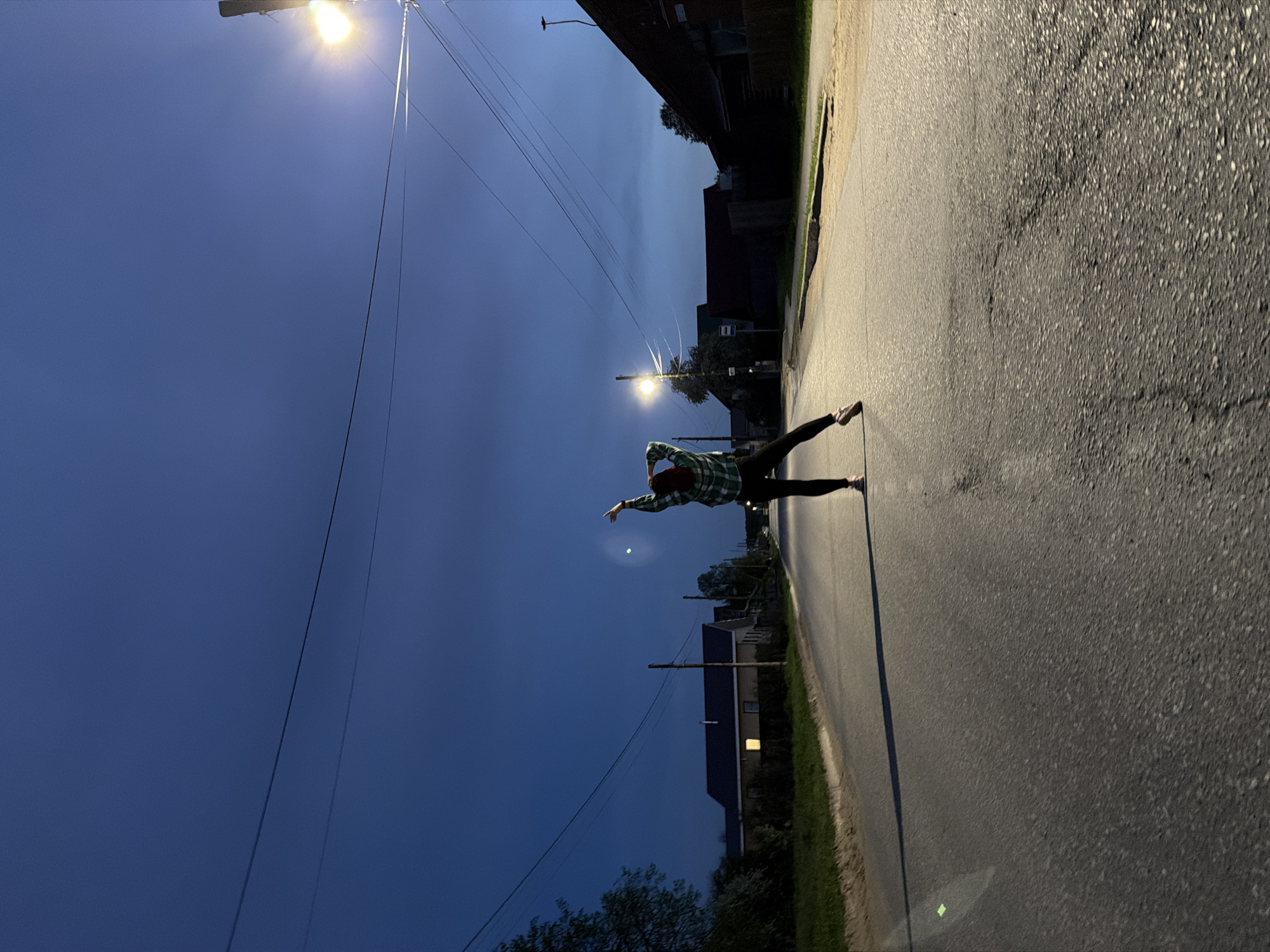 A person wearing dark pants, a green-and-black checkered shirt, and pink shoes is posed on a paved road, arms outstretched to the left. A street lamp is visible in the middle ground, and power lines run across the sky. Buildings and trees are visible along the roadside. The sky is a deep blue. Based on the visible architecture and vegetation, the image could potentially be from a residential area in Chaikovsky, Russia, but without additional context, a precise determination is impossible.