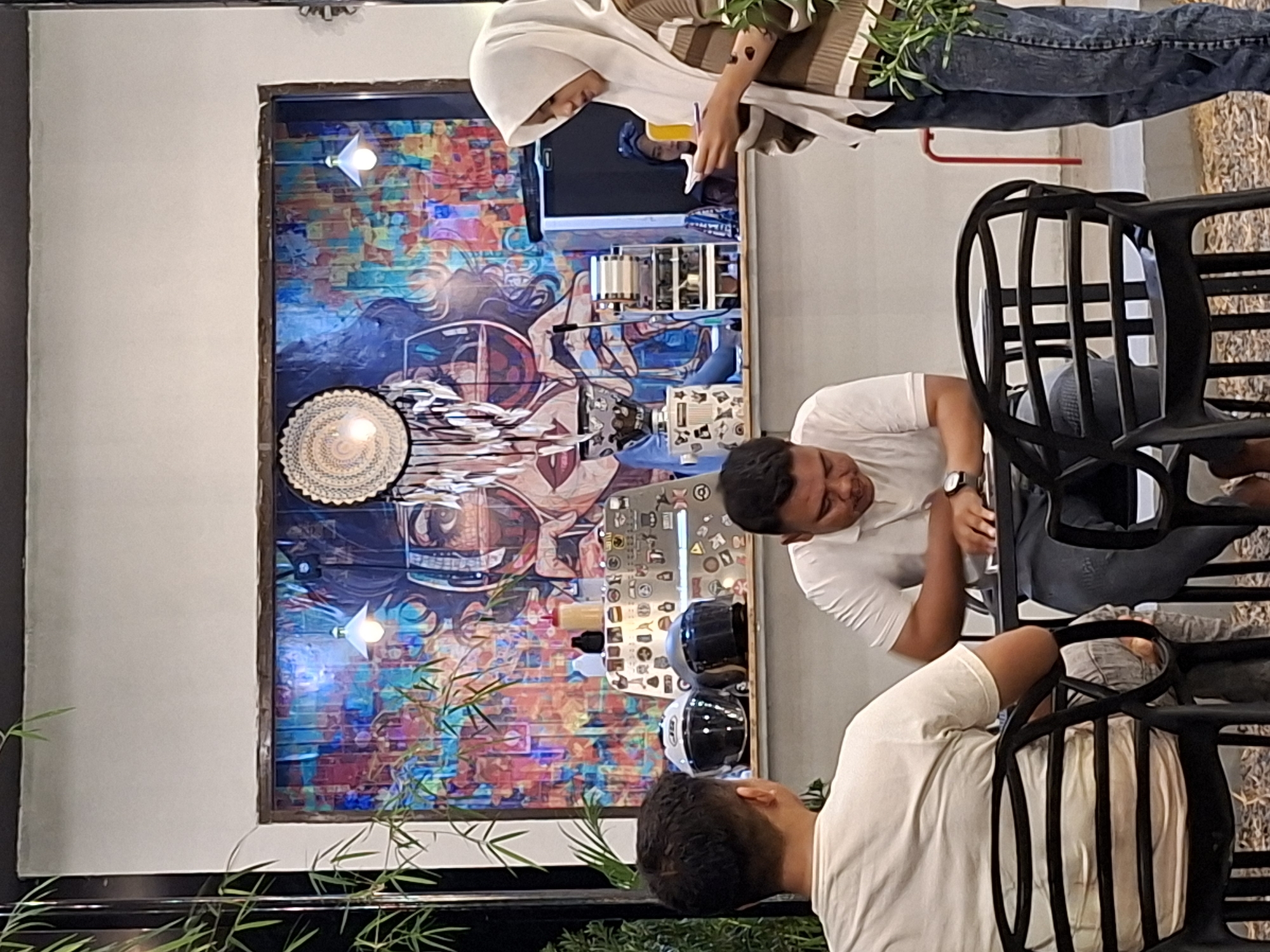 This photograph, taken indoors, likely in Lhokseumawe, Indonesia, shows a group of three people around a table. One person, wearing a white hijab and a striped shirt, is partially visible at the top of the frame, holding something in their hands. To the right is a person in a white shirt, wearing a watch and sitting at the table. The third individual, also in a white shirt, sits facing the second person. Between them are two black chairs. Behind the individuals is a wall decorated with an art piece featuring a collage of colors, images, and objects, including what appears to be motorcycle helmets. Above the artwork are two lights. The setting seems to be a cafe, with a focus on local art.