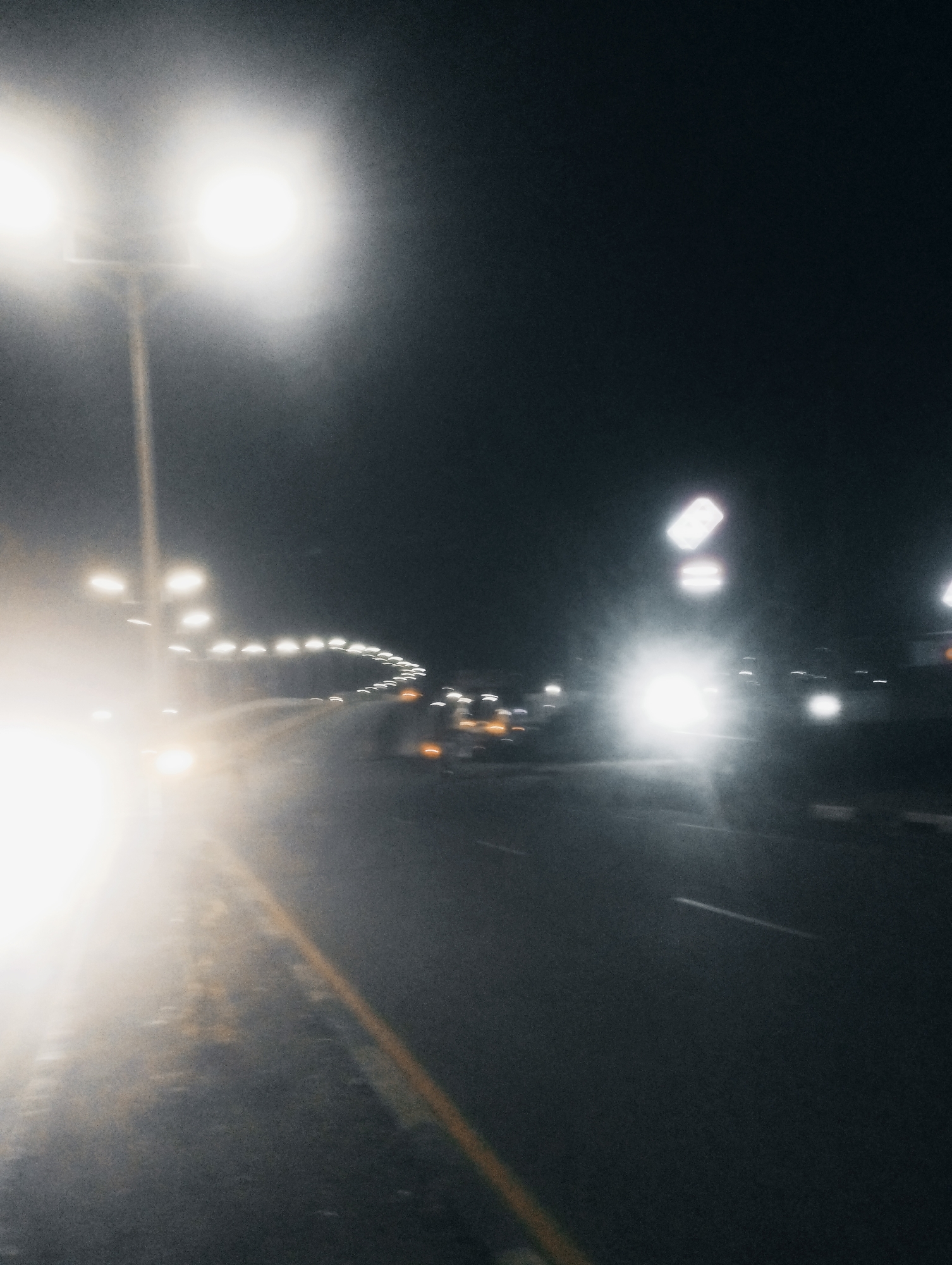 This is a long-exposure photograph of a road at night, likely a highway or major road, given the streetlights and the presence of multiple lanes. The motion blur suggests vehicles are moving. The primary subjects are the streetlights and the road itself. The bright lights of approaching vehicles create streaks and a lens flare, common in night photography with moving lights. The overall scene is dark, with the ambient light coming from artificial sources.
The time of day is clearly night. The atmosphere appears hazy or misty, contributing to the diffused glow of the lights and the softness of the image. This could be due to fog, dust, or light pollution. There are no people clearly visible, but the presence of vehicles implies their activity. The yellow line marking the edge of the road is a notable detail, indicating a well-maintained infrastructure. The background is dominated by the dark night sky, with no visible stars.
The activity taking place is vehicular traffic at night on a road. The impression is of a journey, possibly on a long stretch of road, given the repeating pattern of streetlights. The emotion conveyed is a sense of movement and perhaps isolation or tranquility, depending on the viewer's interpretation of a solitary night journey. There is no visible text in the image. The context of Jos, Nigeria, suggests this could be a road leading into or out of the city, or a highway connecting different parts of the region.
