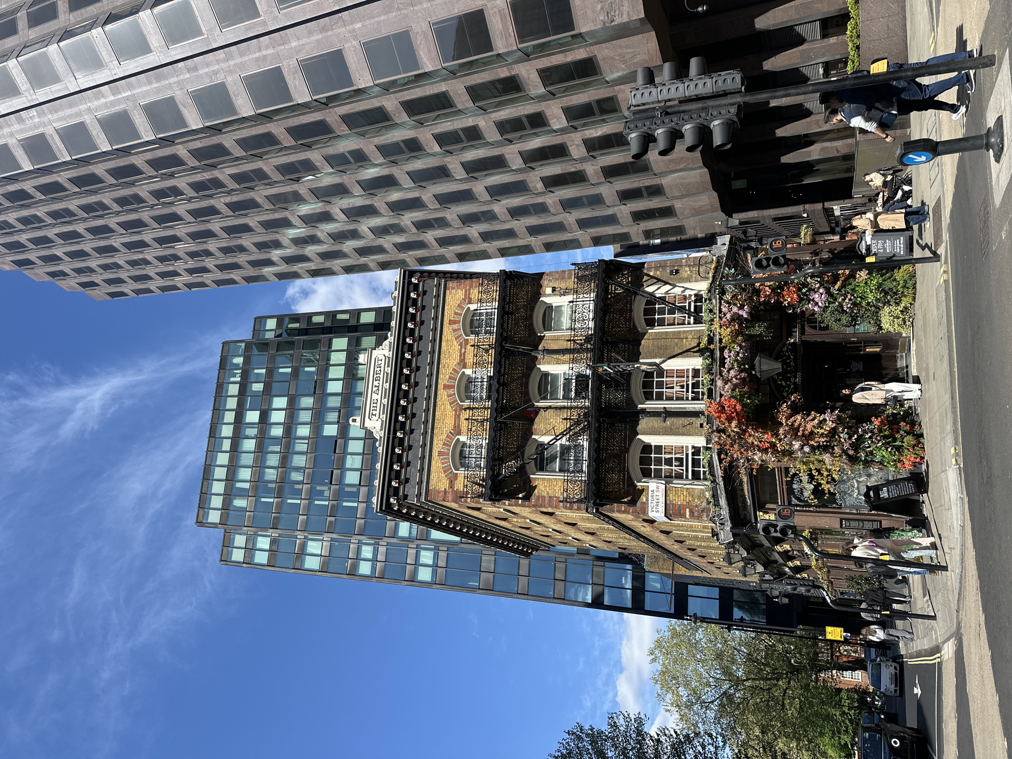 The image depicts a street scene in London, United Kingdom, with a view of a street lined with buildings. On the right, there is a multi-story brick building with a fire escape and a facade adorned with flowers and greenery at the entrance. A sign that reads "The Albert" and a street sign "Victoria Street SW1" are visible. People are walking on the sidewalk in front of the building. A traffic light and street signs are along the sidewalk. Adjacent to the brick building is a modern glass-clad skyscraper. Across the street is another large building with a grid-like pattern of windows. The sky is blue with scattered clouds. A tree with green leaves is in the foreground. A black cab is parked on the street.
