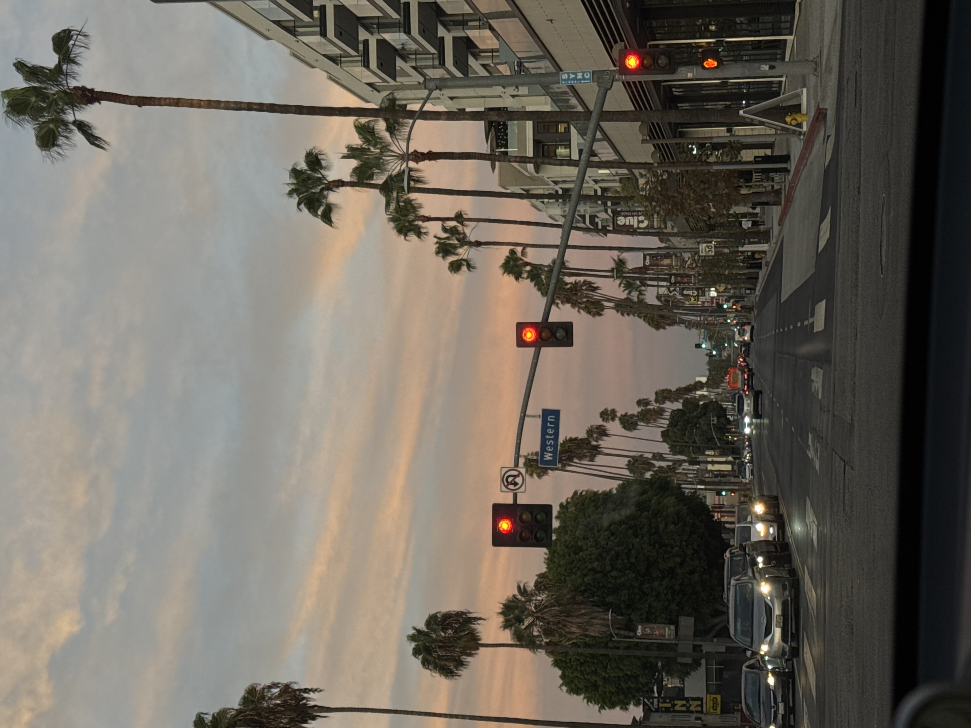 The image depicts a street scene in Los Angeles, California, at dusk. The sky is filled with pastel-colored clouds, suggesting either sunrise or sunset. Palm trees line both sides of the street, with tall, slender ones dominating the right side, interspersed with shorter, fuller ones on the left.
The street is an urban thoroughfare with multiple lanes of traffic. Several cars are visible, some in motion and some stopped at a traffic light. The traffic lights themselves are illuminated, with one showing red and another yellow. Street signs are present, including a "Western" street sign and a speed limit sign of "30". Buildings of modern architecture, featuring balconies and glass facades, line the right side of the street. On the left side, there are glimpses of storefronts and trees. The perspective is from inside a vehicle, looking forward down the street. The overall impression is that of a busy urban environment bathed in the soft light of the approaching evening.