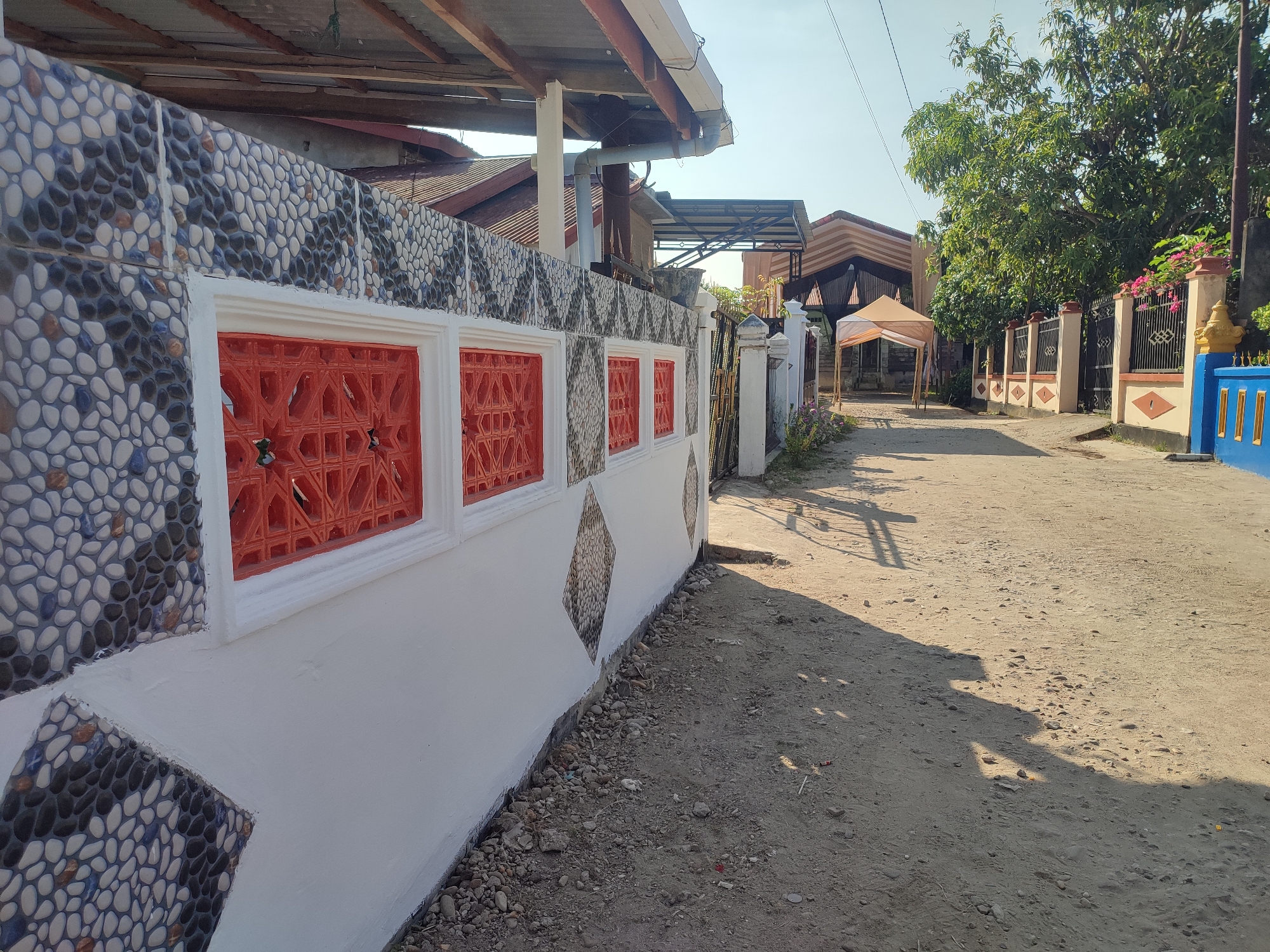 The image depicts a dusty, unpaved street in Gampongjawa, Indonesia. On the left, a low wall with decorative stone tiling and red latticework is visible, partially shaded by an overhanging roof. The wall transitions into a gate and fence extending along the right side of the street. A beige tent-like structure is set up further down the road, casting shadows. The street is lined with houses and lush green trees in the background. The sky is clear and blue.