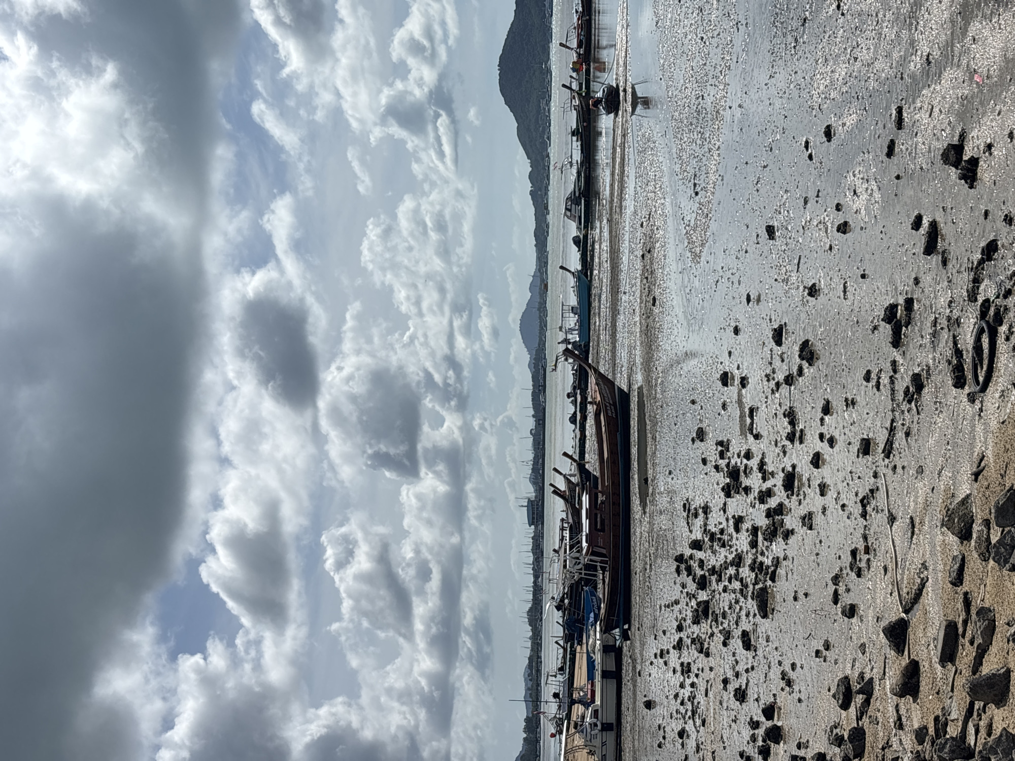 The image depicts a low tide scene at Rawai Beach, Thailand. Numerous long-tail boats are beached on the exposed muddy shore, some pulled up higher than others. A wooden fishing boat, brown and tan with a covered cabin, is prominently featured in the foreground. The shore is littered with rocks of various sizes, and tidal pools reflect the cloudy sky. In the distance, a calm body of water separates the beach from a hazy coastline with rolling hills and islands. The sky is filled with dramatic, textured clouds, suggesting a partly cloudy day. The overall atmosphere is one of a quiet, working waterfront during low tide. There are no people visible in the image.