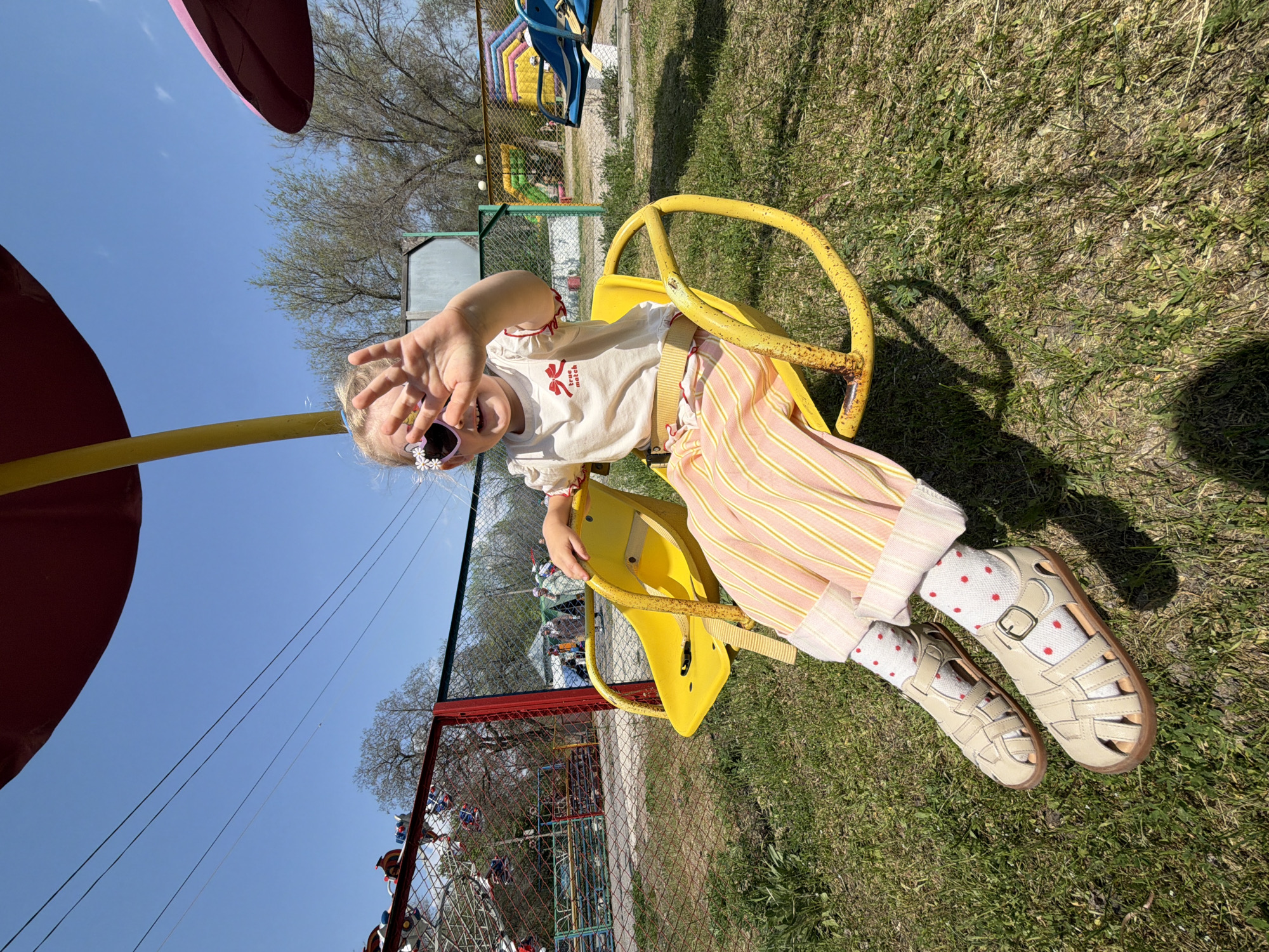 A young girl sits on a yellow merry-go-round in Kamyshin, Russia. She has on sunglasses shaped like flowers, white socks with red polka dots, and tan sandals. She is wearing striped pants and a white shirt. The merry-go-round is on a green lawn. A blue sky is overhead, with trees and a fence in the background.
