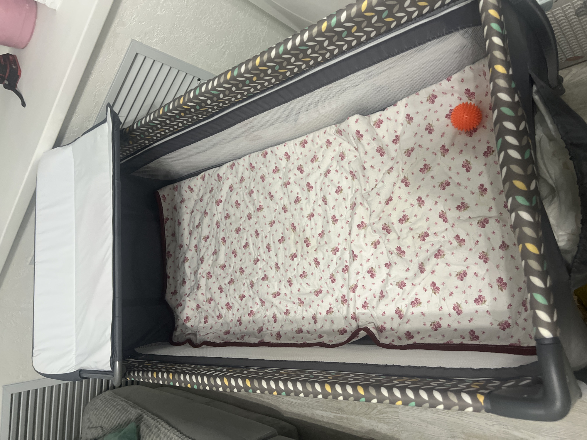 The image shows a portable crib with a white canopy and a floral-patterned sheet. An orange spiky ball is visible on the sheet. The crib appears to be in a room with a light-colored wall and a wooden floor. The location is identified as Surgut, Russia, and the scene is depicted with a neutral, objective tone, focusing on observable details without interpretation or speculation.