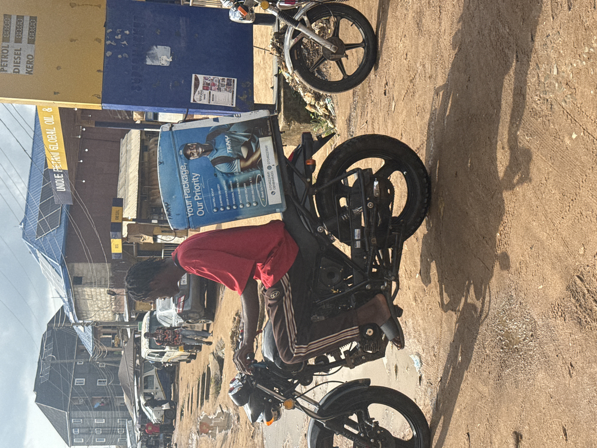 The image depicts a street scene in Ihiagwa, Nigeria. A young man is seated on a black motorcycle with a delivery box attached to the back, bearing the logo of "TENY" and the slogan "Your Package, Our Priority." The box also displays a phone number and lists delivery services.
The man wears a red shirt and dark pants with white stripes. He has his hands on the handlebars and appears ready to drive. He is wearing sandals. The street is unpaved and shows signs of wear.
In the background, various buildings, power lines, and vehicles, including white vans and cars, are visible. There are also other people on the street. A fuel station sign is seen indicating the availability of petrol, diesel, and kerosene. A banner of a fuel company is also visible.