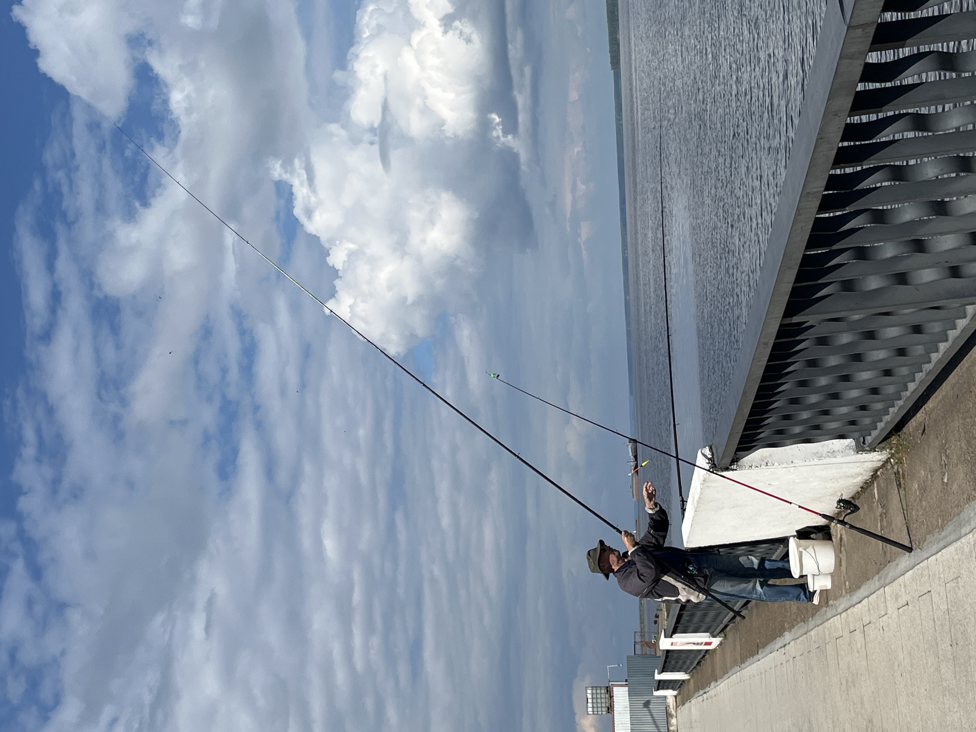 The image depicts a solitary fisherman on a concrete embankment or pier, engaged in the activity of fishing on a large body of water, likely the Kama River in Chaikovsky, Russia. The scene is set under a bright, cloudy sky.
The main subject is a man, seen from the side, wearing a dark jacket, blue jeans, and a wide-brimmed hat. He is holding one fishing rod with both hands, appearing focused on his line or adjusting his tackle. Beside him, two other fishing rods are propped up against the railing of the embankment, equipped with reels and visible floats (one green, one yellow). Two white buckets are placed on the ground near the propped rods, possibly for bait or to hold caught fish.
The embankment features a concrete surface and a distinctive metal railing with horizontal, wavy slats on its inner side, extending along the edge of the water. The water body stretches widely into the distance under the overcast sky, with subtle ripples visible on its surface. A faint shoreline or landmass can be seen on the horizon. The sky is largely covered with fluffy cumulus clouds, indicating a daytime setting, likely mid-day or afternoon, with bright but diffused light.
In the far distance on the left side of the frame, some industrial-looking structures are vaguely visible. There is no discernible text in the image. The overall impression is one of a peaceful and focused fishing outing.