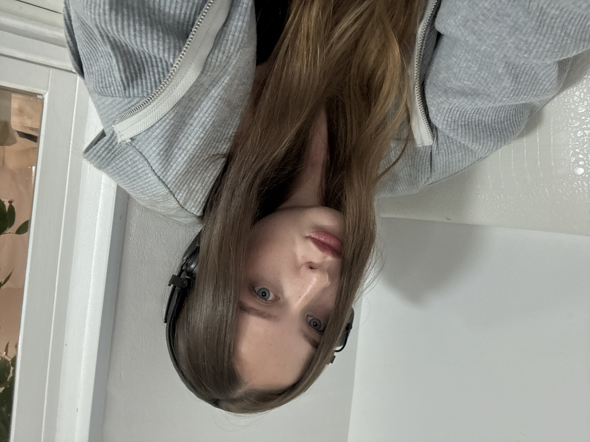 A young woman with long brown hair and blue eyes is pictured from the chest up, wearing black headphones and a grey zippered hoodie. She is looking directly at the camera with a neutral expression. The background consists of a white wall and a glimpse of a window on the right side of the frame, with some greenery visible outside. The lighting is soft, suggesting an indoor environment. There is no indication of her location in Chaikovsky, Russia.