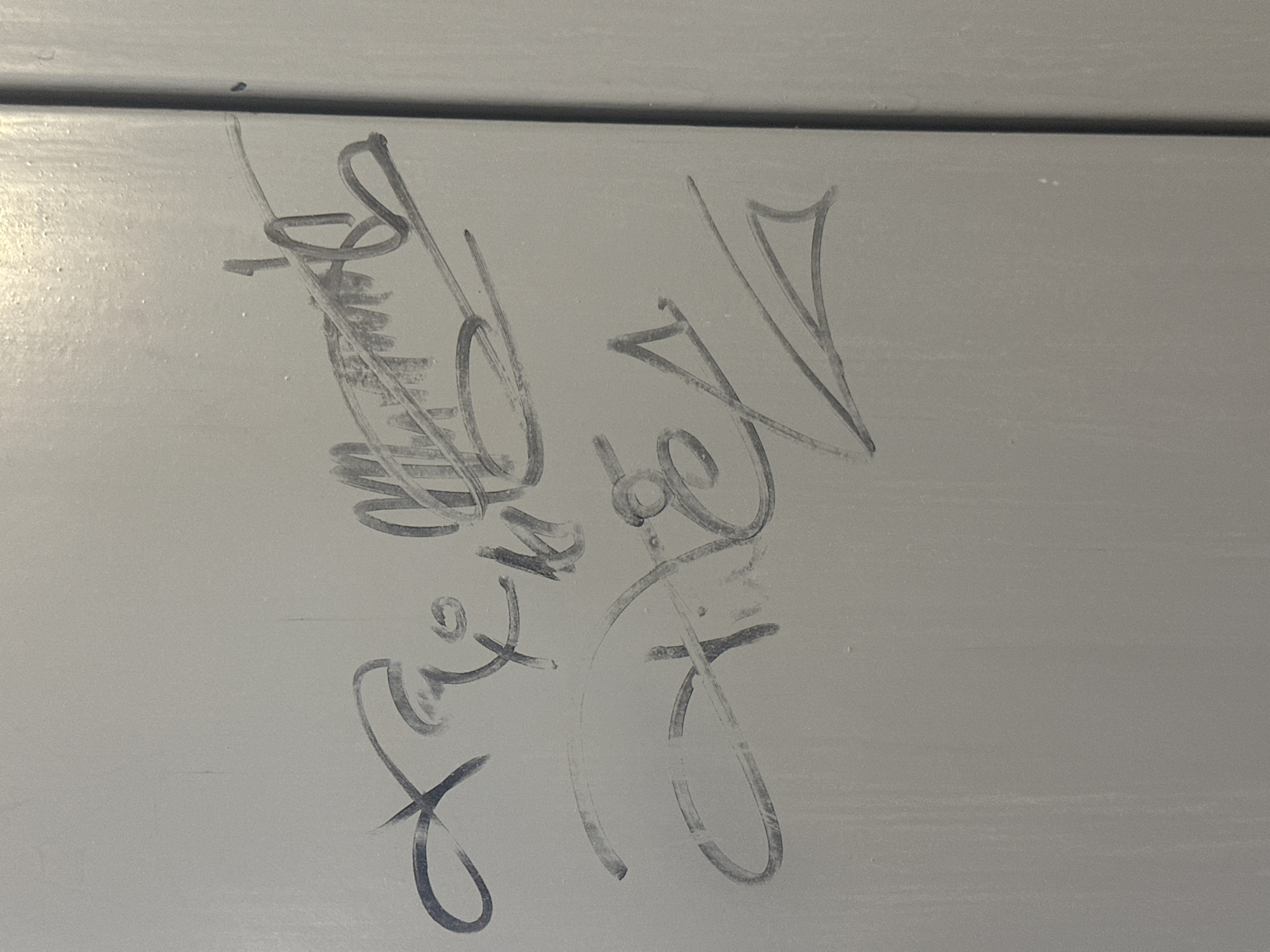 A grey, textured surface, possibly a wall or door, is marked with two sets of dark grey graffiti or tags. The graffiti appears to be handwritten with a marker or similar writing instrument. The first tag is located in the upper half of the image, characterized by a series of interconnected loops, curves, and a central scribbled area. The second tag is situated below the first, also composed of curvilinear strokes, and some angular lines that form a pointed shape. The lighting suggests the image was taken indoors or in a shaded outdoor area, with a slight sheen on the surface. The background is a uniform light grey. The graffiti is the sole subject of the image. There is no information available about the specific location in Uzhhorod, Ukraine, or any individuals present.