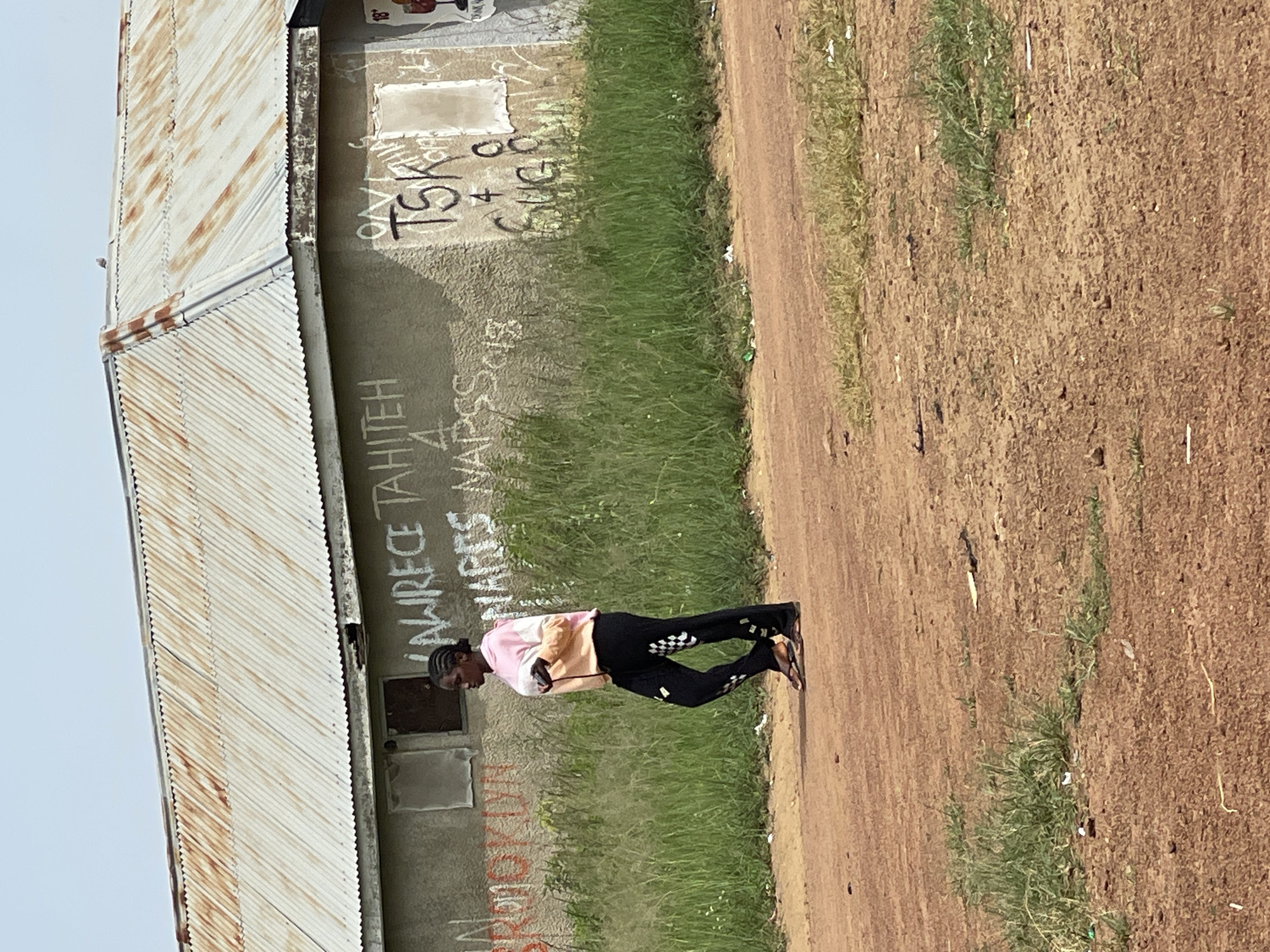 A young Black woman walks across a dirt road in front of a building with a corrugated metal roof and graffiti on its concrete walls in Durumi, Nigeria. The woman is wearing a pink and white long-sleeved shirt and black pants with a checkered pattern on the sides. She is carrying a white object in her left hand, which may be a phone. The building has two visible windows with dark panes, and the roof shows signs of rust. The graffiti on the wall includes white lettering that appears to say "LAWRENCE TAHITEH" and "NAPSS NAPSS 2018," as well as "TSK" and other markings. The ground in front of the building is covered in dry grass and scattered debris. The sky is overcast.