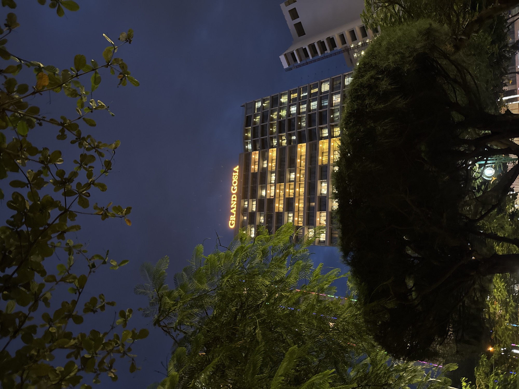 This image depicts the Grand Gosia hotel in Nha Trang, Vietnam, at night. The hotel is a tall, multi-story building illuminated by warm lights in its windows, creating a grid pattern against the dark blue sky. The hotel's name, "GRAND GOSIA," is prominently displayed in glowing orange letters at the top of the building. In the foreground, lush green trees with dense foliage obscure parts of the hotel and the sky. Some of the trees have leaves that appear to be illuminated from within or are reflecting light. There are also some colorful lights, possibly from decorations, visible on the trees, creating streaks of light. A portion of another building, lighter in color with visible structural elements, is visible to the right of the Grand Gosia hotel. The overall scene suggests a warm, urban evening setting.