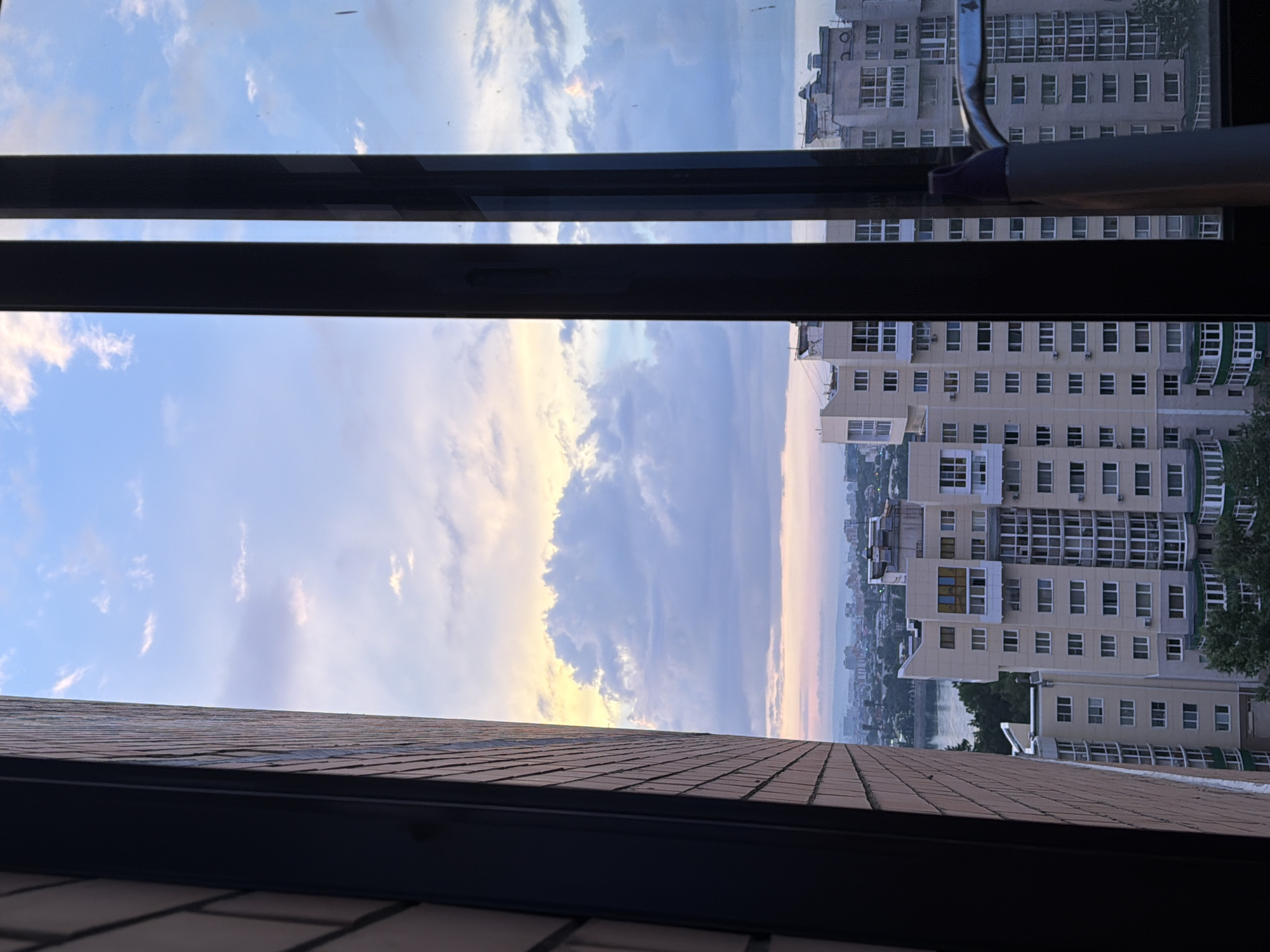 The image is a photograph taken from a window overlooking a city at dusk. The sky is filled with dramatic clouds, ranging in color from pale blue to soft pinks and yellows, indicating the sun is low on the horizon. In the foreground, two vertical black window frames divide the view. To the left, a brick wall is visible, showing a rough texture and a light brown color. Beyond the window frames, a cityscape unfolds. Tall, multi-story residential buildings dominate the middle ground, characterized by numerous windows and balconies. Some buildings are light-colored, possibly beige or white, while others appear to be a mix of colors. A body of water, likely a river or lake, is visible in the distance, reflecting the twilight sky. The background shows a hazy outline of more distant buildings and hills, suggesting a sprawling urban area. No people or specific objects like vehicles are clearly discernible. The overall impression is a view of a city during the transitional period between day and night. Given the distinctive architecture of the buildings and the presence of a large body of water within a city, this scene could plausibly be a view from an apartment window in Irkutsk, Russia, a city located on the shore of Lake Baikal.