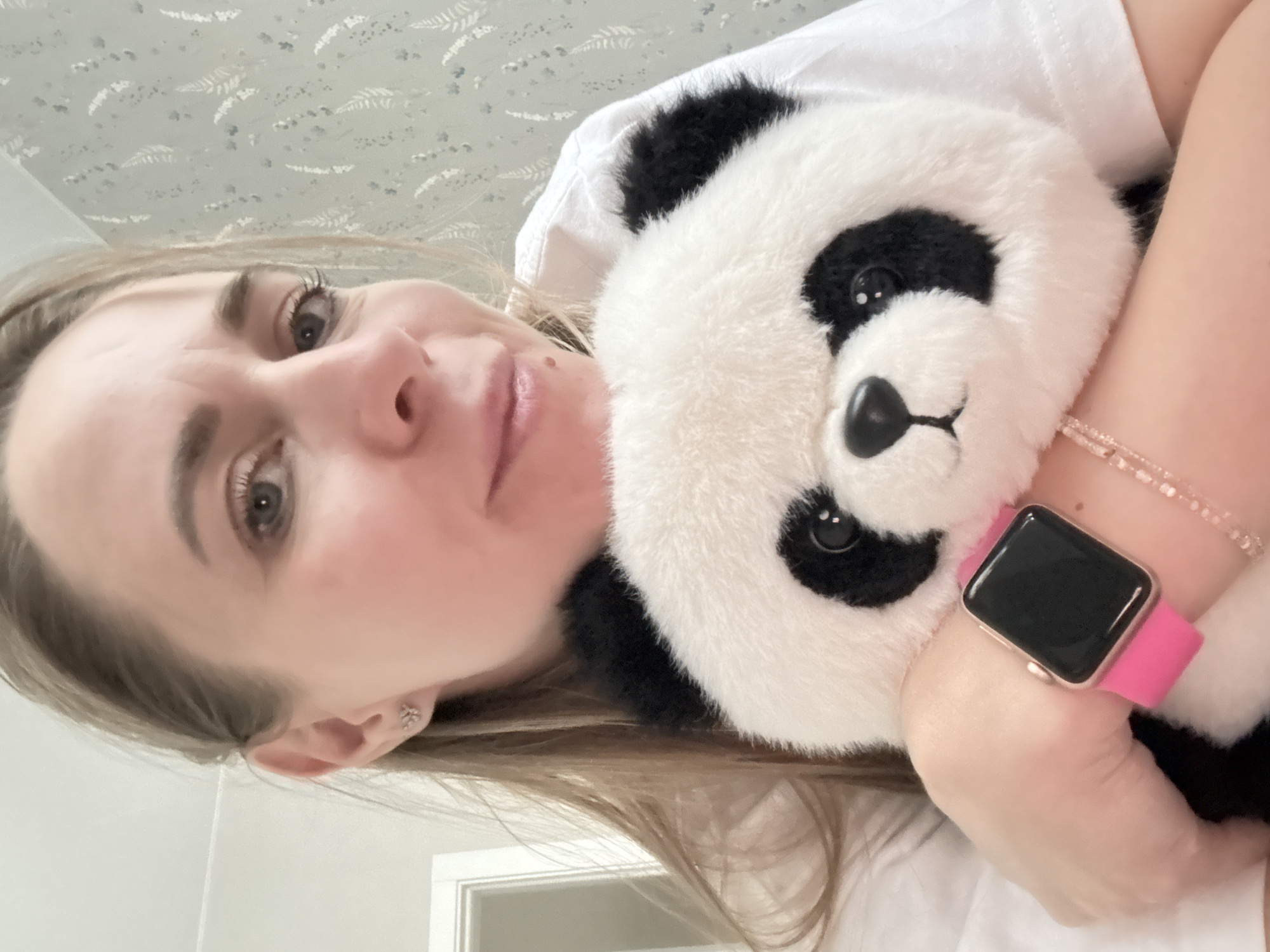 A woman is holding a stuffed panda bear. She is wearing a pink-banded smartwatch on her left wrist and a white t-shirt. Her blonde hair is parted and falls to one side. The background shows a wall with a light-colored patterned wallpaper and a white door frame. The location is not identifiable as Chaikovsky, Russia based on visual information.