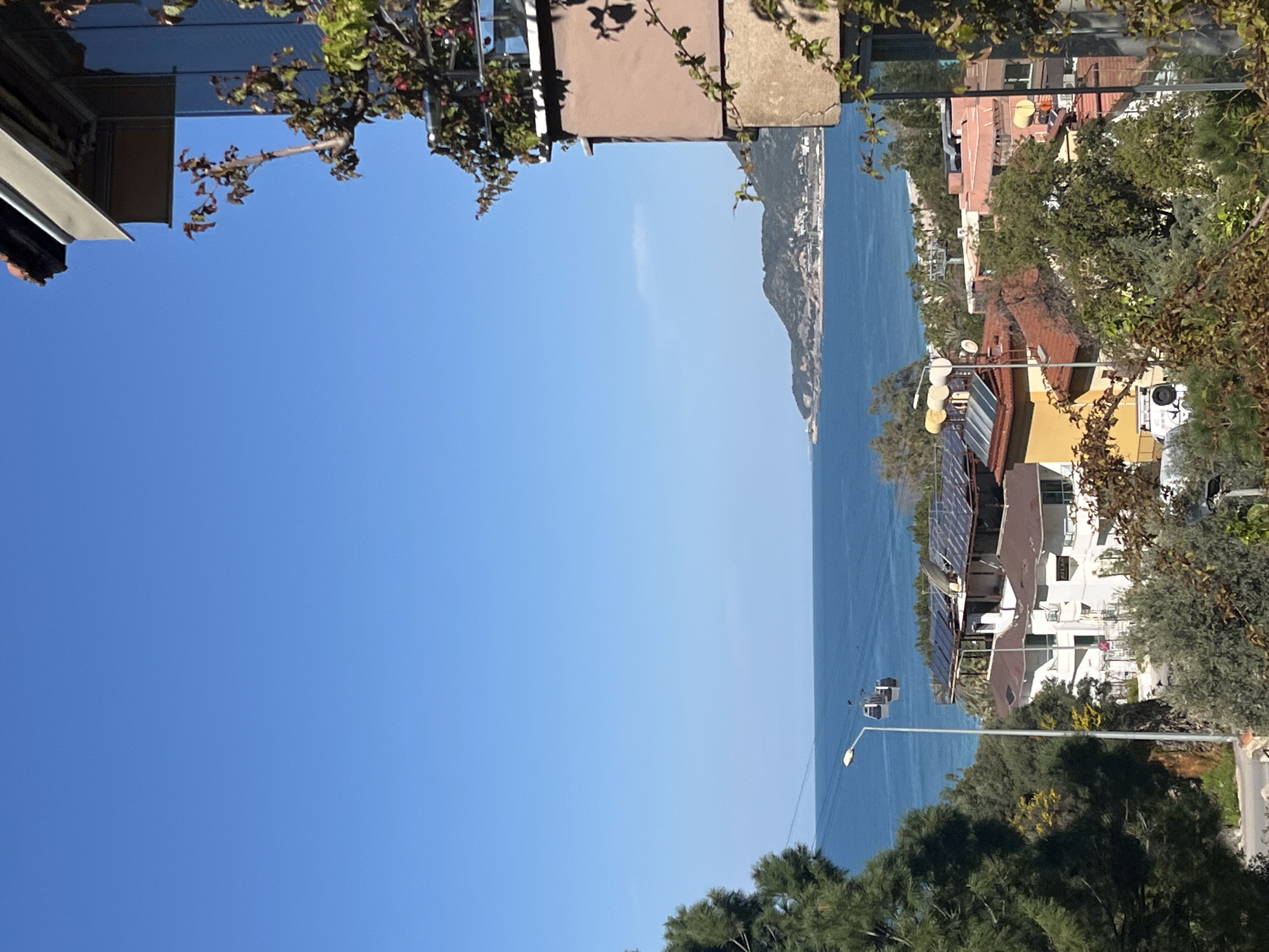 The image shows a view from a high vantage point, possibly a balcony, overlooking the sea in Alanya, Turkey. In the background, there's a mountainous peninsula extending into the sea. Buildings with red-tiled roofs and white facades are visible, some equipped with solar panels. A cable car system operates over the water. The foreground includes foliage and architectural elements from the viewing point. The sky is clear and blue.