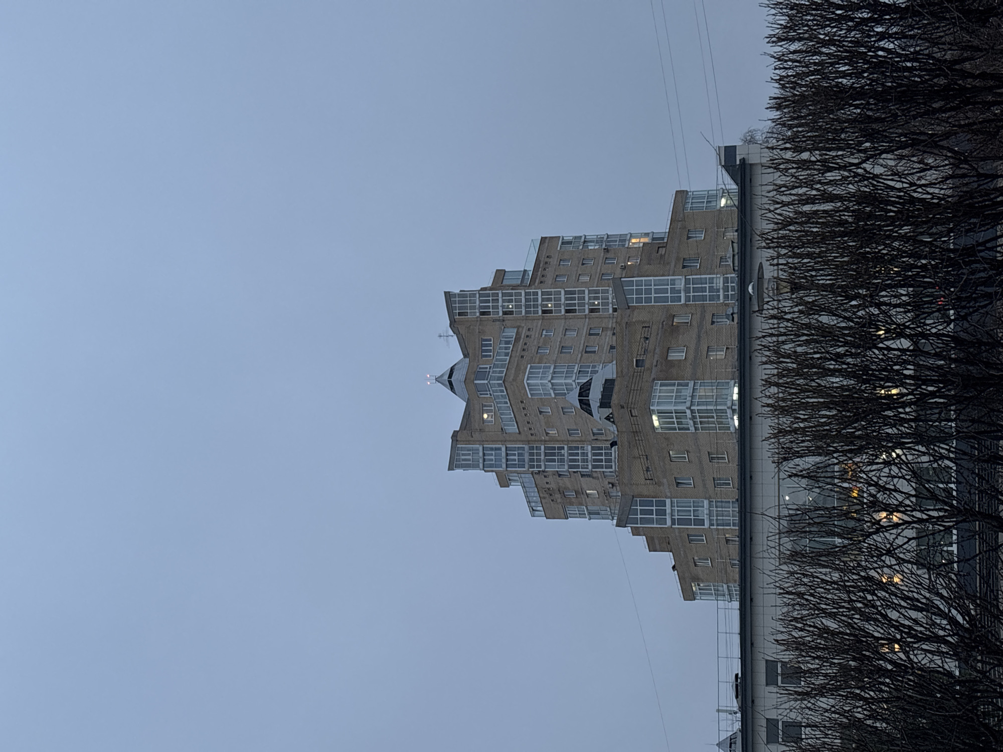 The image shows a multi-story building with a unique terraced design against a grey sky. The building is constructed of light-colored brick or stone and features many windows. Part of another building is visible to the right, along with the bare branches of trees in the foreground. The location appears to be Perm, Russia, due to the architectural style of the building. There are no people visible in the image. The scene is muted, with an overcast sky.