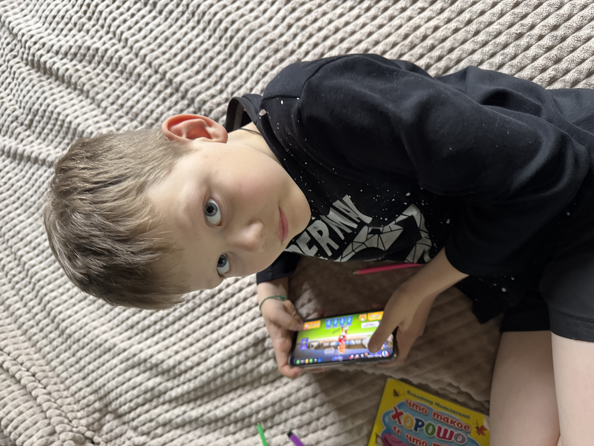 A young child is lying on a textured, light-brown surface, possibly a bed or couch. The child is fair-skinned with short blonde hair and blue eyes. They are wearing a black hooded sweatshirt with white splatter designs and black shorts. The child is holding a smartphone in their hands, with a colorful game displayed on the screen. A pink pencil lies near the phone, and a yellow children's book is visible in the lower right corner. Part of the book's text is visible in Russian. The location is in Ферма, Russia.