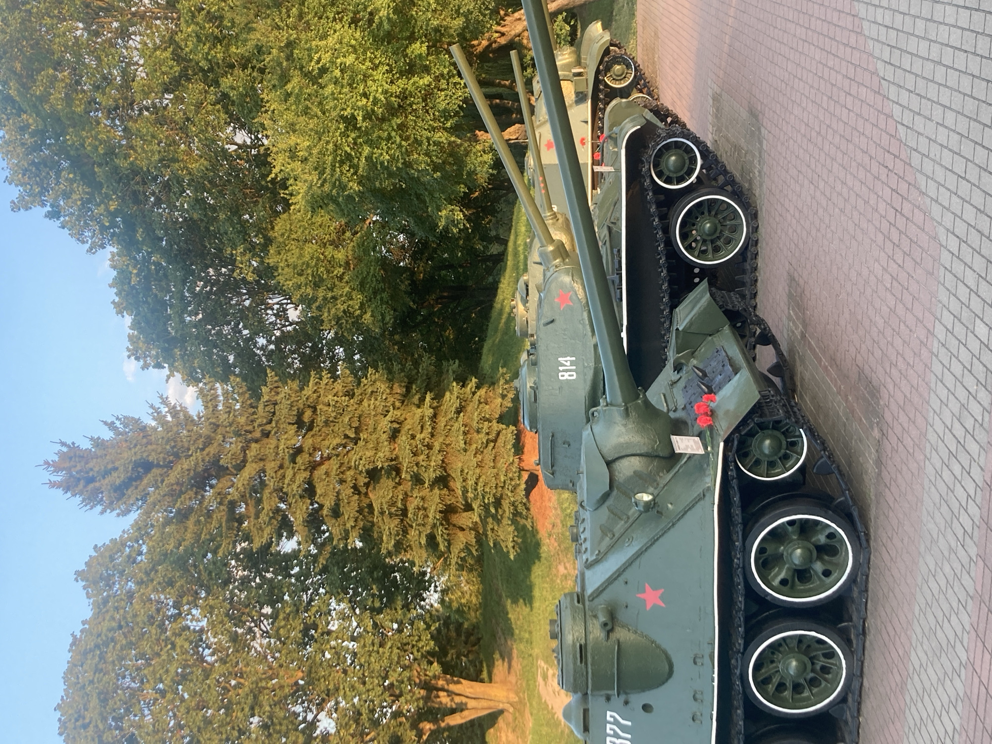 The image shows a row of several green tanks with red stars displayed on a paved walkway in front of a grassy incline with trees. The tanks are oriented from left to right, with the closest tank partially obscuring the ones behind it. The front tank has the number "377" visible on its side. The second tank from the front has the number "814" on its side. The tanks appear to be on display, likely at a memorial or museum. The background consists of mature trees and a clear blue sky with some white clouds. The paving stones are a mix of red and grey. There are no people visible in the image. This scene is likely located at the Brest Hero Fortress Memorial Complex in Brest, Belarus, a prominent historical site.
