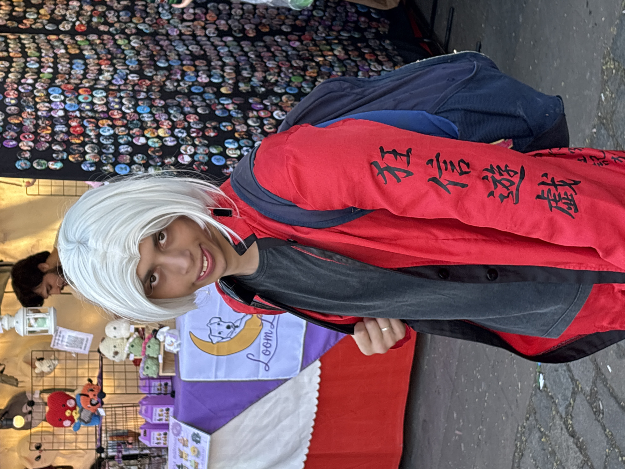 A person with short, white, spiky hair and a light-skinned complexion is wearing a red jacket over a dark grey t-shirt and a dark backpack. The person is looking at the camera and smiling. The jacket has black Japanese characters embroidered on the sleeve. Behind the person, to the left, is a vendor's booth with various handmade items for sale, including plush toys and small decorative objects. To the right, a wall is covered in hundreds of small, circular pins or badges. The ground appears to be cobblestone. The setting suggests an outdoor market or street fair.