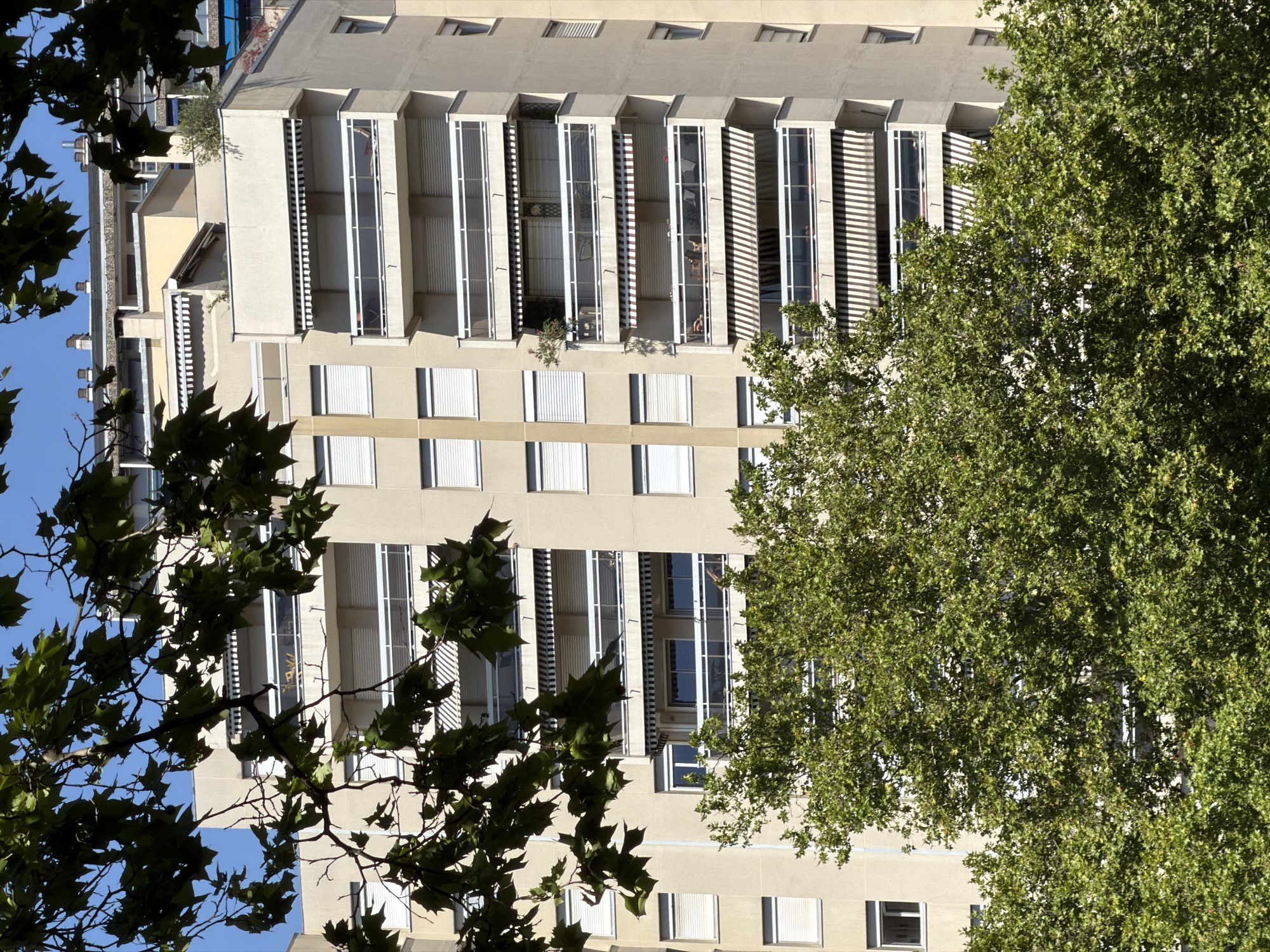 The image depicts a multi-story apartment building, likely located in the city of Lyon, France, as suggested by the architecture and general ambiance. The building is primarily light beige in color and features a series of balconies with glass railings and white horizontal accents. Many of the balconies are equipped with retractable awnings, some striped black and white, and others solid.
On the balconies, various objects are visible, including lounge chairs, small tables, and potted plants. Through the windows and balcony doors, glimpses of interior spaces can be seen, suggesting residential apartments. The building is partially obscured by lush green foliage from trees in the foreground, with leaves and branches silhouetted against the building and the clear blue sky. The leaves appear to be from a deciduous tree, possibly a plane tree, common in urban environments.
In the background, other buildings of varying heights and architectural styles are visible, some with rooftop chimneys or other structures. The overall scene suggests a warm, sunny day in an urban setting, with a blend of natural and man-made elements. There are no people clearly discernible in the image, and no specific actions or interactions are depicted. The focus is on the architecture of the building and its integration with the surrounding greenery.