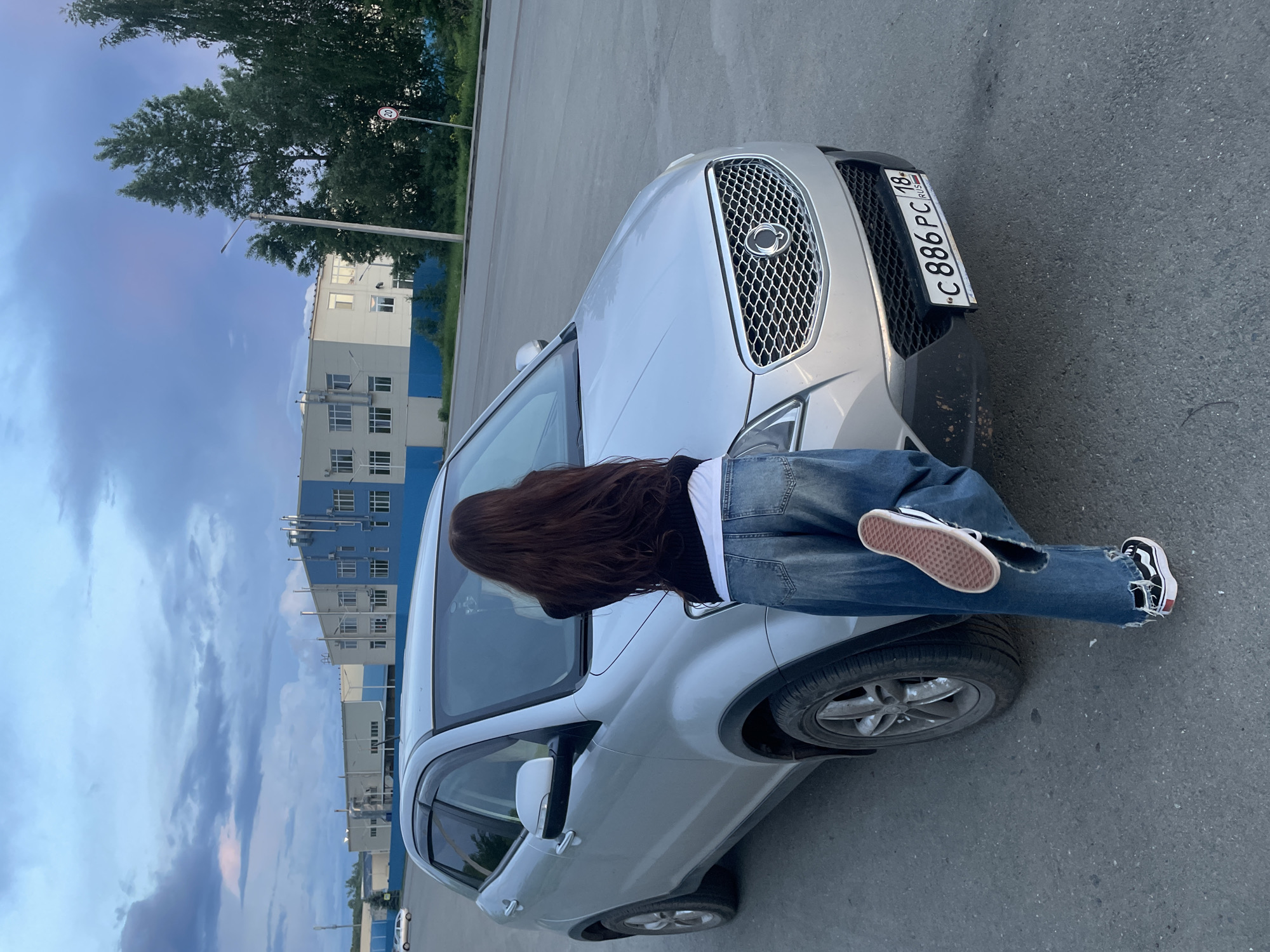 The image, captured in portrait orientation but displayed rotated 90 degrees counter-clockwise, depicts an outdoor scene featuring a person and a silver SUV. The setting is a paved area, likely a street or parking lot, against a backdrop of trees, modern buildings, and a twilight sky.
A person with long, dark brown hair is the central human subject, seen from behind as they lean into the open driver's side window of the SUV. They are dressed in a black top, a white undershirt, and distinctive ripped blue jeans paired with black and white sneakers featuring reddish-brown soles. Their posture suggests they are either entering, exiting, or interacting with the car's interior.
The silver SUV, possibly a SsangYong based on its grille design, is prominently featured, with its front grill and emblem clearly visible. The Russian license plate, "C 886 PC 18 RUS," is affixed to its front bumper.
In the background to the left, green trees stand tall, accompanied by a street light and a round speed limit sign displaying "20". To the right, multi-story buildings with contrasting light grey and blue sections and numerous windows suggest a commercial or industrial area. The sky above shows hues of blue and light purple, indicative of late afternoon or early evening. The overall impression is one of a calm, clear day at dusk in an urban or suburban environment.