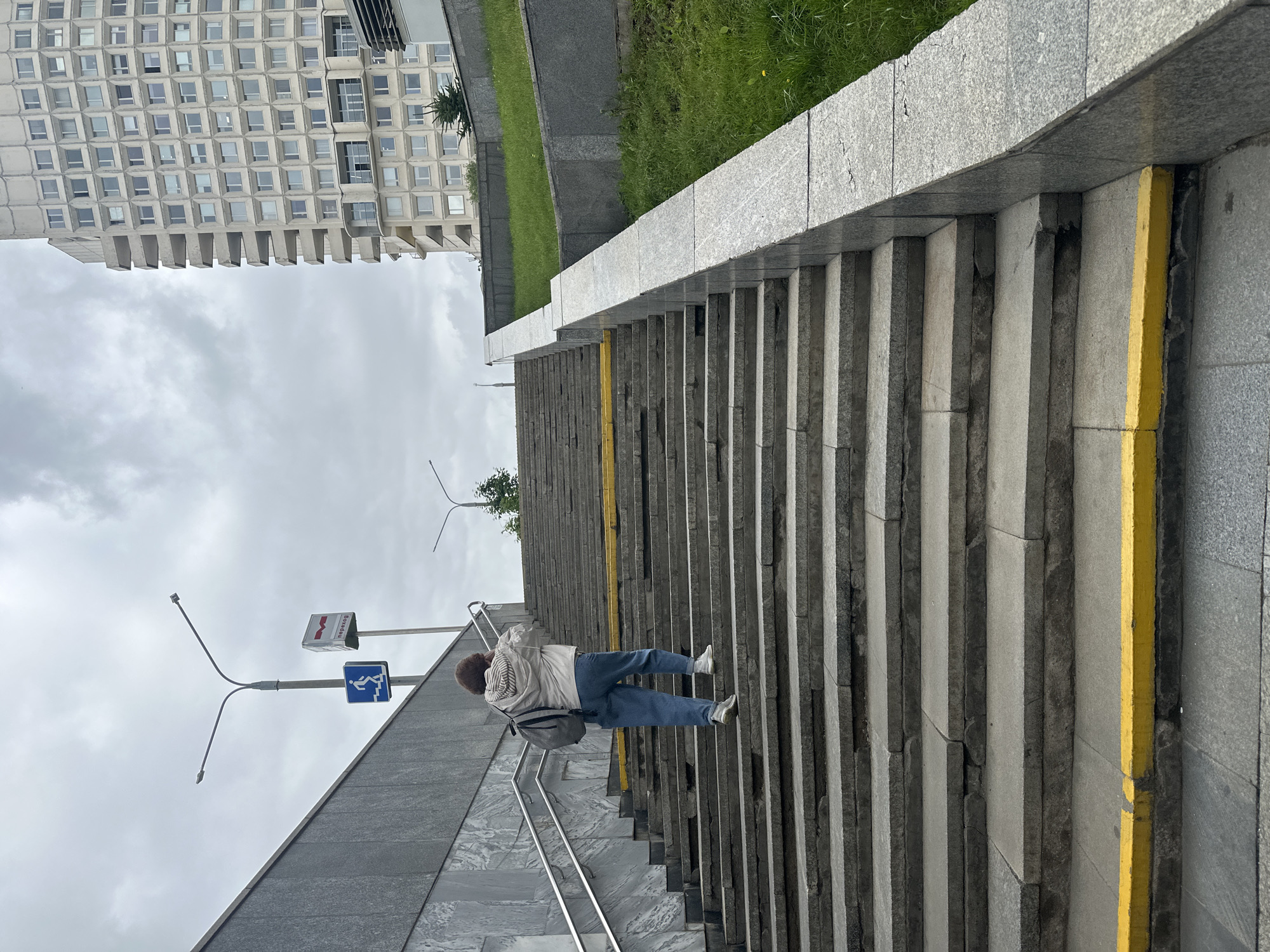 A person with dark curly hair, wearing a light-colored hooded jacket and blue jeans, walks up a wide, stone staircase. They are carrying a grey backpack over their left shoulder. The staircase has a yellow stripe on the edge of several steps. To the left of the stairs, a grey granite wall curves upwards, with a silver handrail. A blue sign with a white icon of a person going up stairs is mounted on a pole. Further up the pole is a rectangular sign with red and white lettering. A tall, modern building with many windows and balconies dominates the background on the right side of the image. Green grass covers a slope to the right of the stairs. The sky is overcast with grey clouds. This image was taken in Minsk, Belarus, likely near a metro station given the signage.