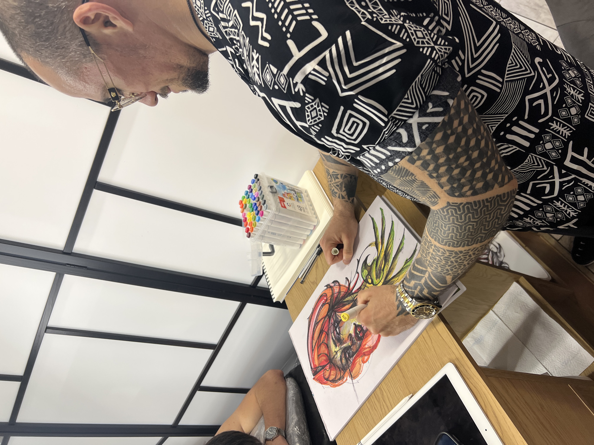 An overhead shot shows a man working on a drawing at a light wooden desk. The man has a closely cropped haircut and beard and wears glasses, a black and white patterned shirt, and a gold and silver wristwatch. His arms are heavily tattooed. He is coloring a drawing with a yellow marker. The drawing is of a stylized creature, rendered in red, orange, yellow, and green. To the left of the drawing is a container of markers. A white tablet and stylus are on the desk. A person's head and shoulder are visible in the lower left of the frame. Behind the desk is a wall composed of rectangular white panels separated by black bars. The location is Sofia, Bulgaria.
