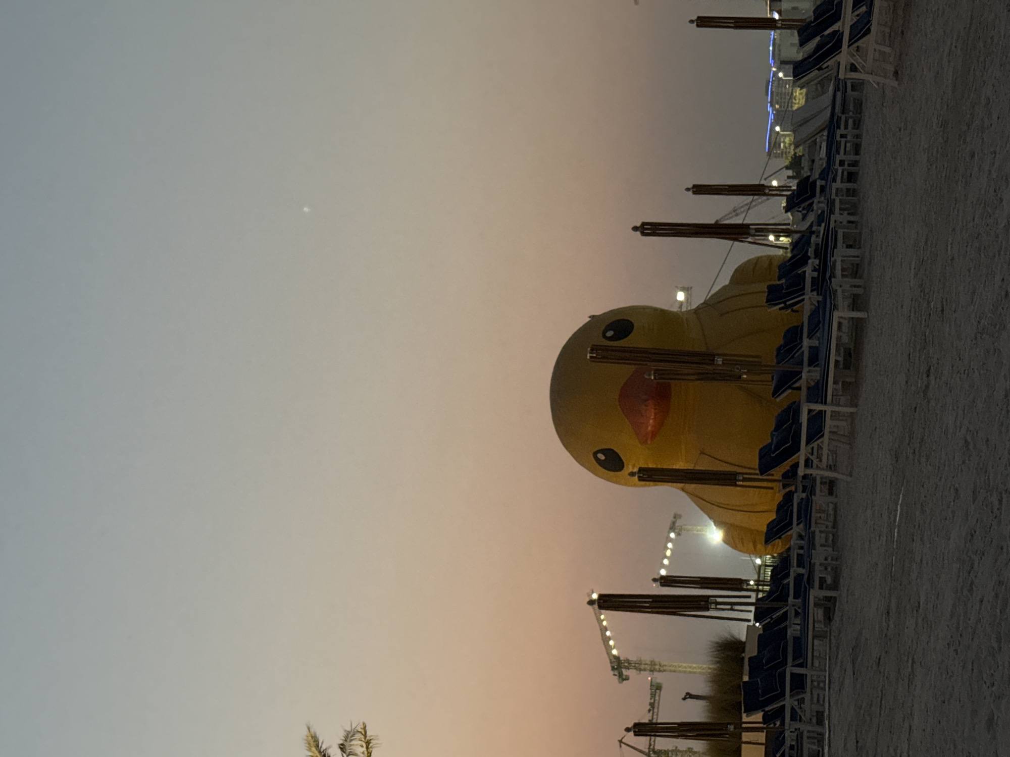An inflatable yellow duck is the central subject of this image, located on a sandy beach in the city of Unknown, United Arab Emirates. The duck is large and positioned in the foreground, facing the viewer. Behind the duck, rows of empty lounge chairs with blue cushions are arranged on the sand, some with umbrellas. In the background, construction cranes are visible against a pale orange and blue gradient sky, suggesting ongoing development. The scene appears to be at dusk or dawn, with soft lighting and a serene atmosphere.