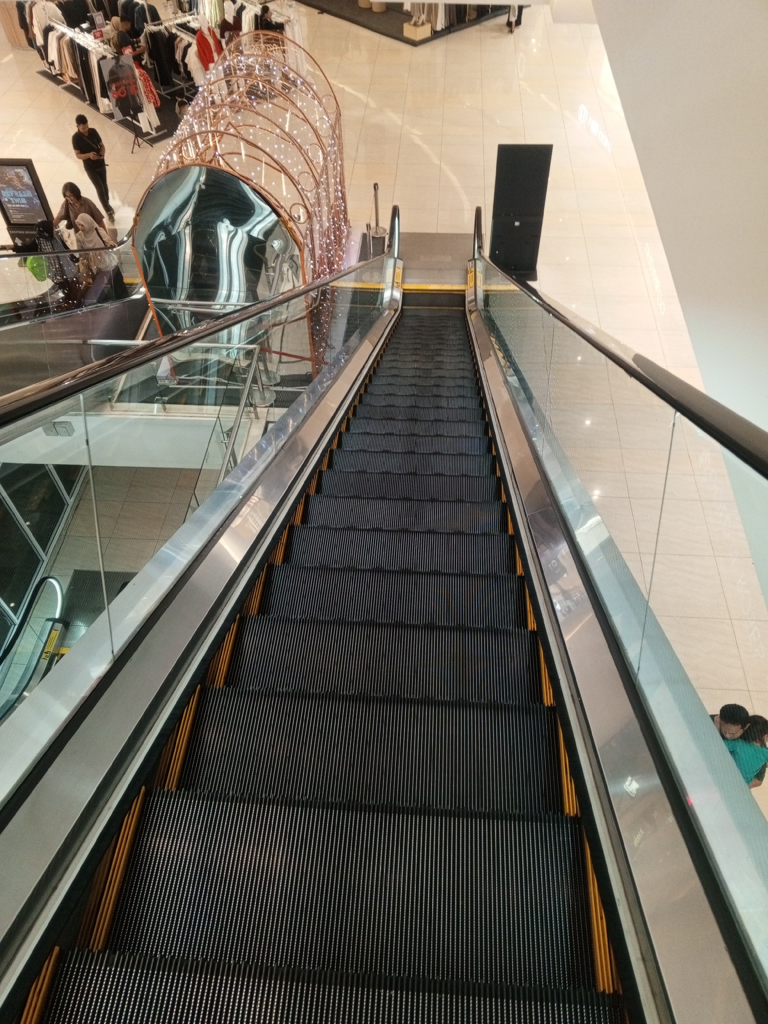 This image captures an upward view of an escalator in a brightly lit indoor shopping mall, likely located in Cibinong, Indonesia. The escalator's dark steps are bordered by reflective metallic sides and clear glass railings. In the upper left background, racks of clothing are visible in a retail store. A decorative, illuminated structure, possibly a temporary display, arches over the escalator's upper landing. Several individuals are present: a man wearing a black t-shirt stands near the clothing racks, while a woman in a brown top and a person in a hijab are on the escalator's upper section. Further down the escalator, two people, one wearing a teal shirt, are partially visible near the bottom. The floor of the mall is tiled in a light, neutral tone.